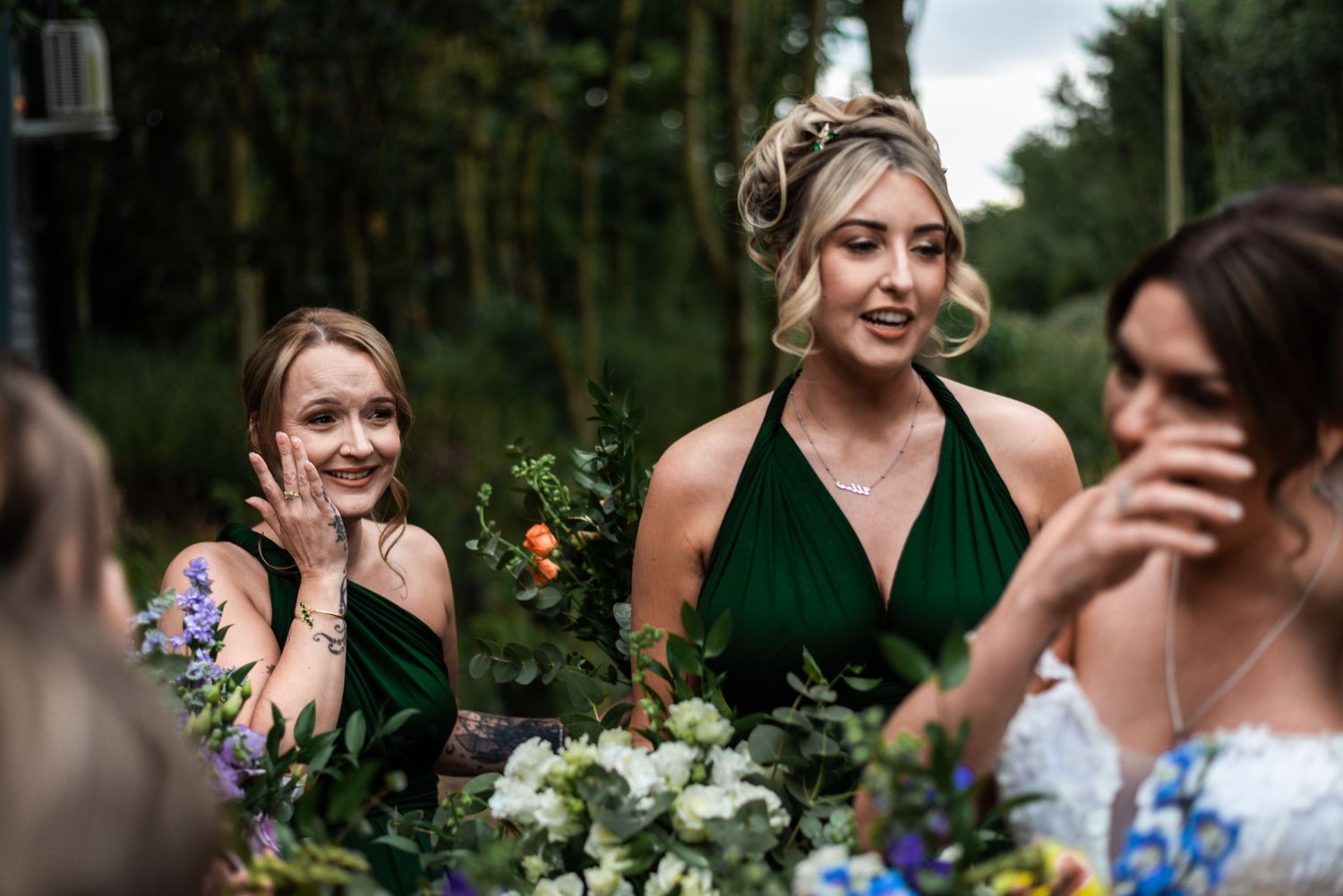 Bridesmaids in emerald green dresses laughing and holding floral bouquets at an outdoor wedding ceremony.