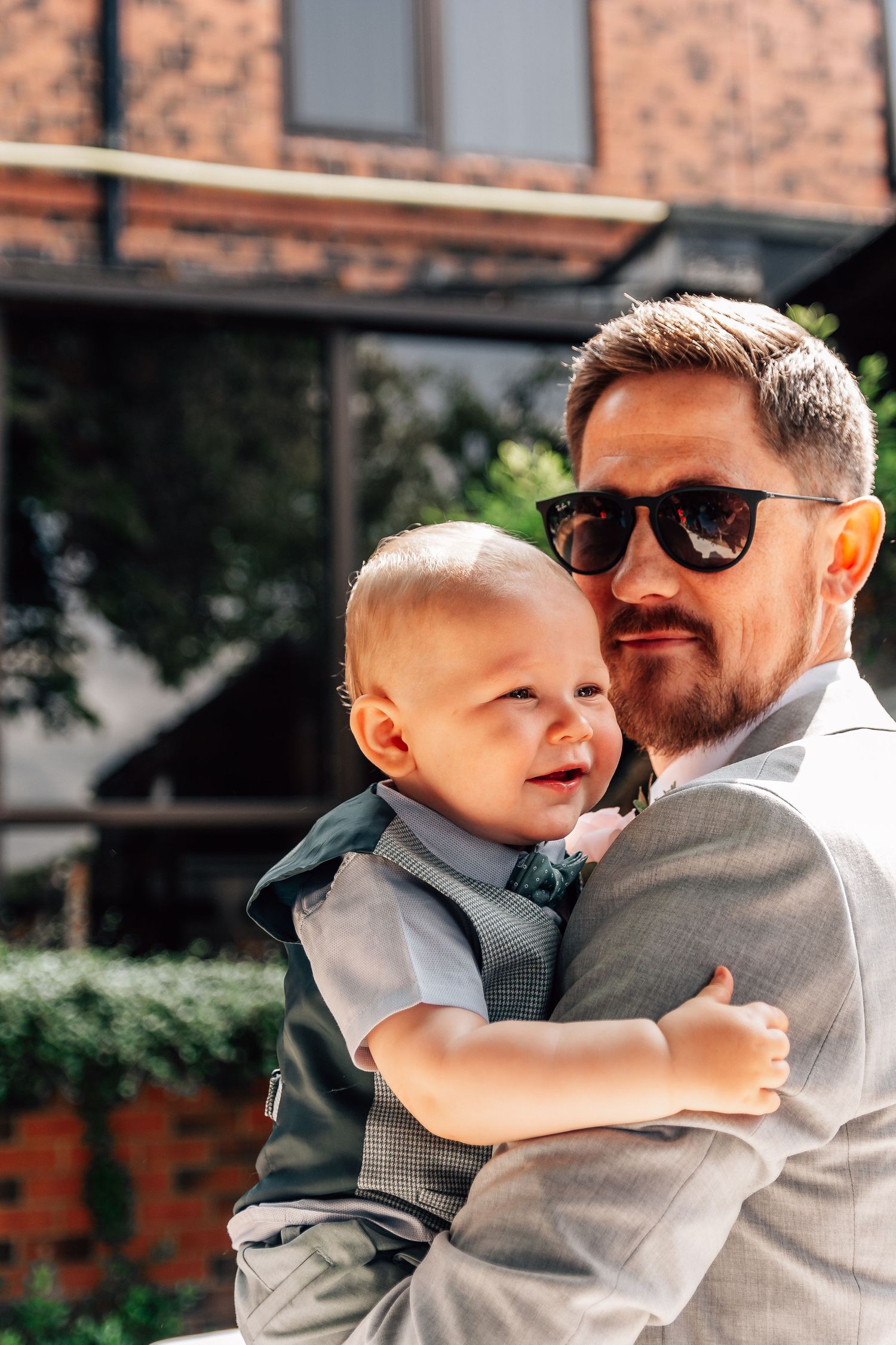 Father in grey suit holding baby boy at outdoor wedding venue.