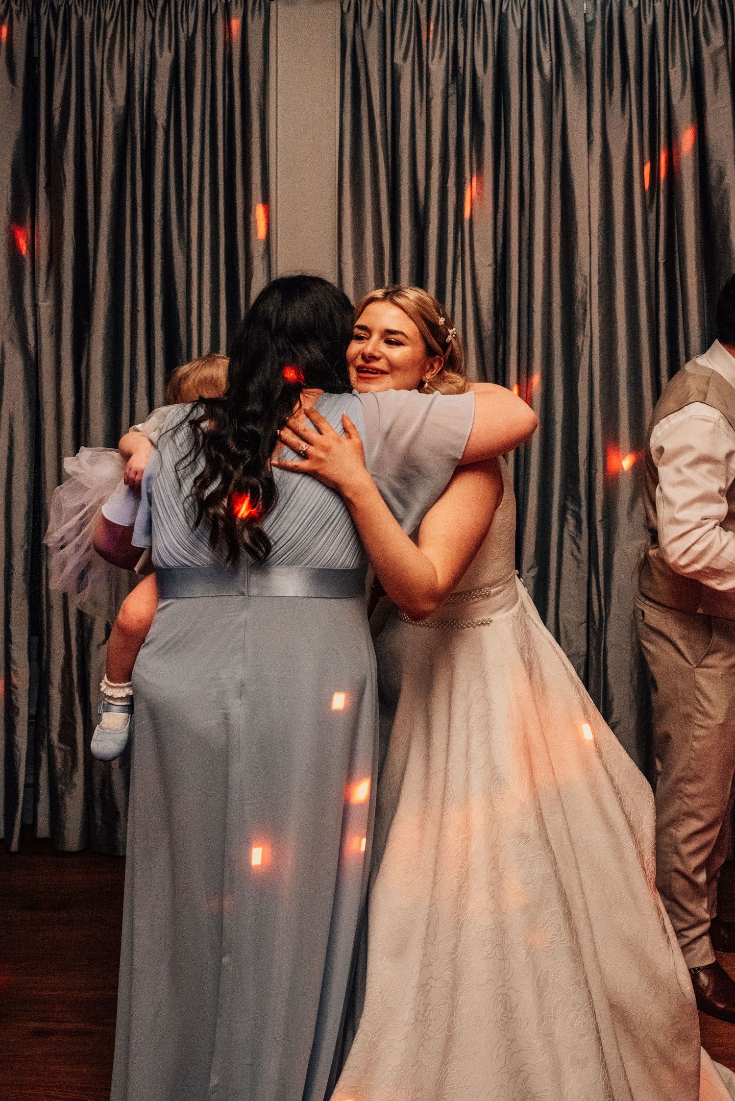 Bride in white lace gown shares warm hug with bridesmaid in blue dress on wedding dance floor.