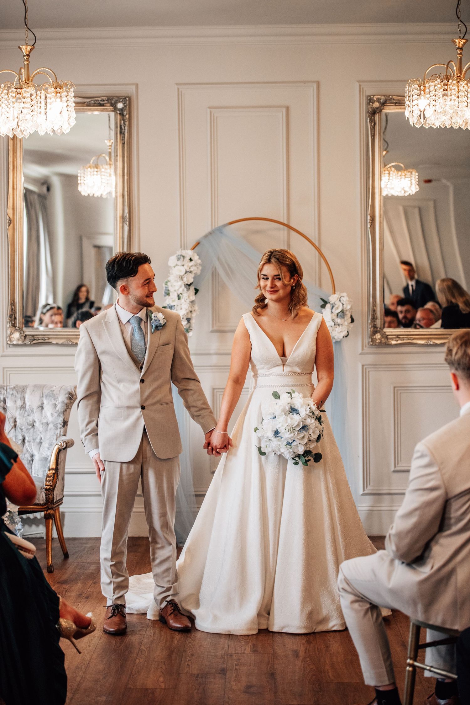 Bride and groom holding hands at elegant wedding ceremony with floral arch, chandeliers, and gold mirrors.