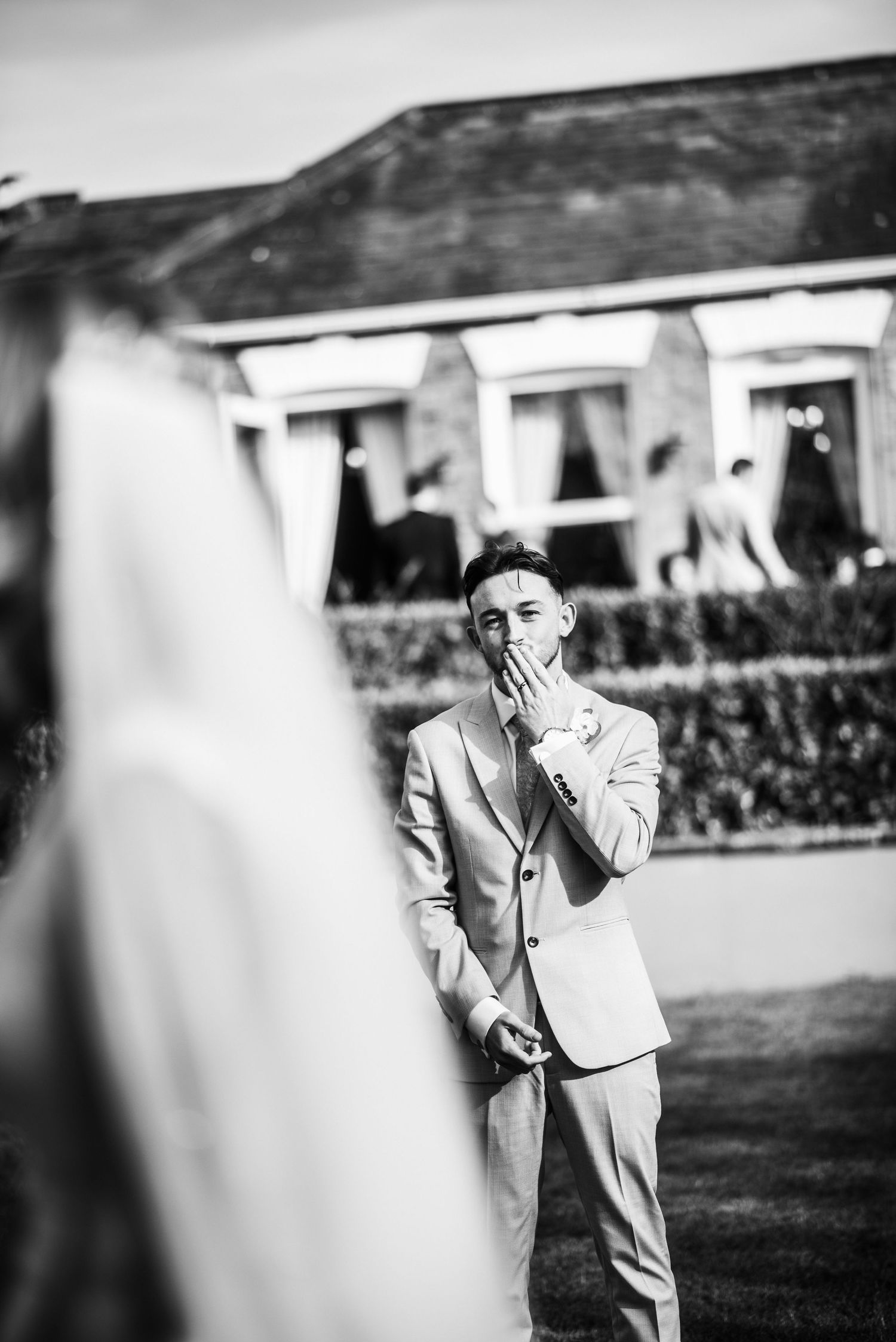 Emotional groom in suit covers mouth in awe seeing bride in black and white wedding photo outdoors.