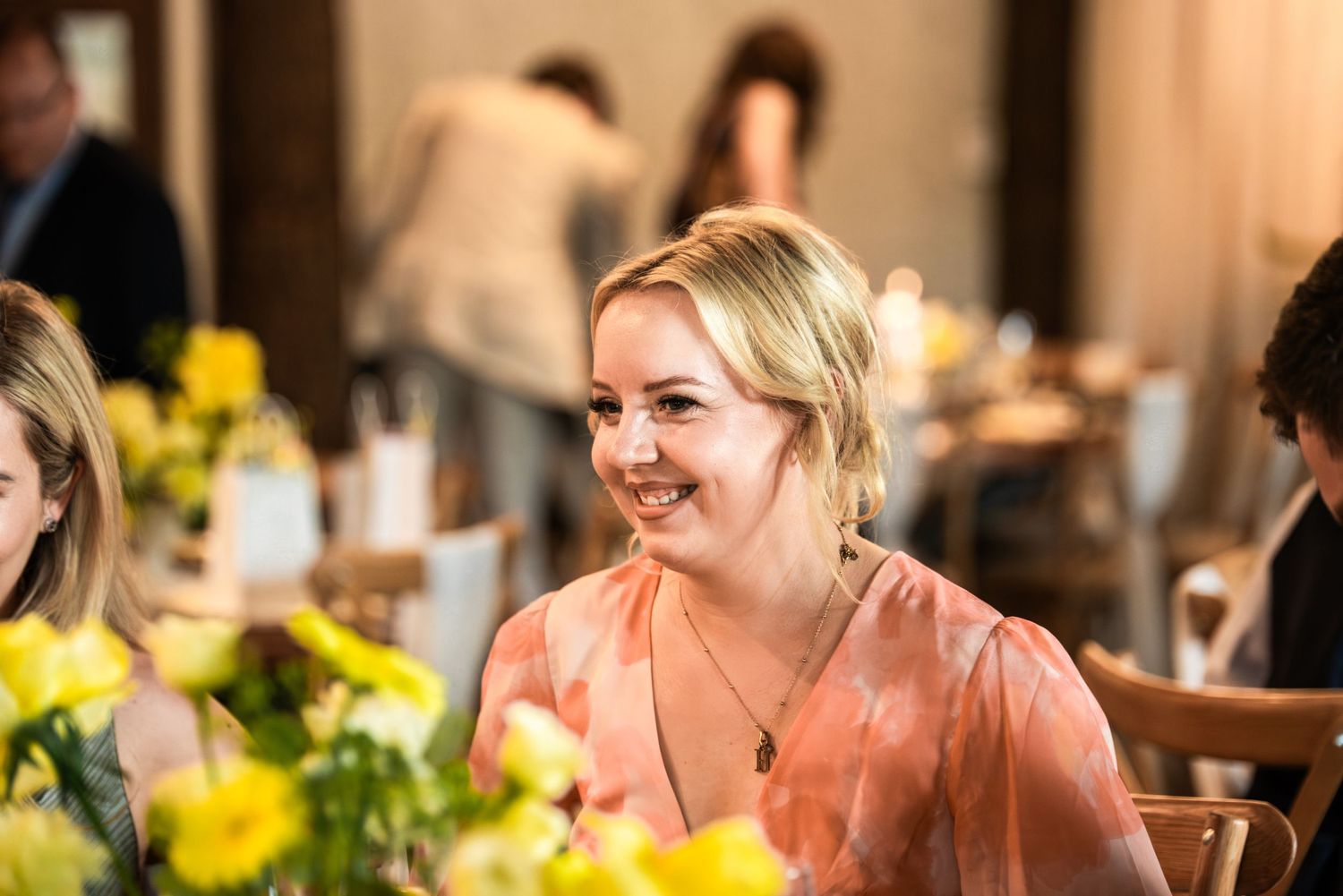 Smiling blonde woman in pink floral dress at elegant dinner event with yellow flowers.