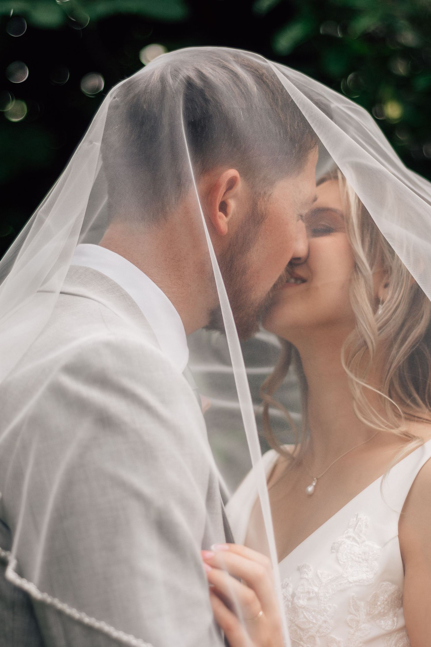 Romantic bride and groom sharing a tender kiss beneath a flowing white wedding veil outdoors.