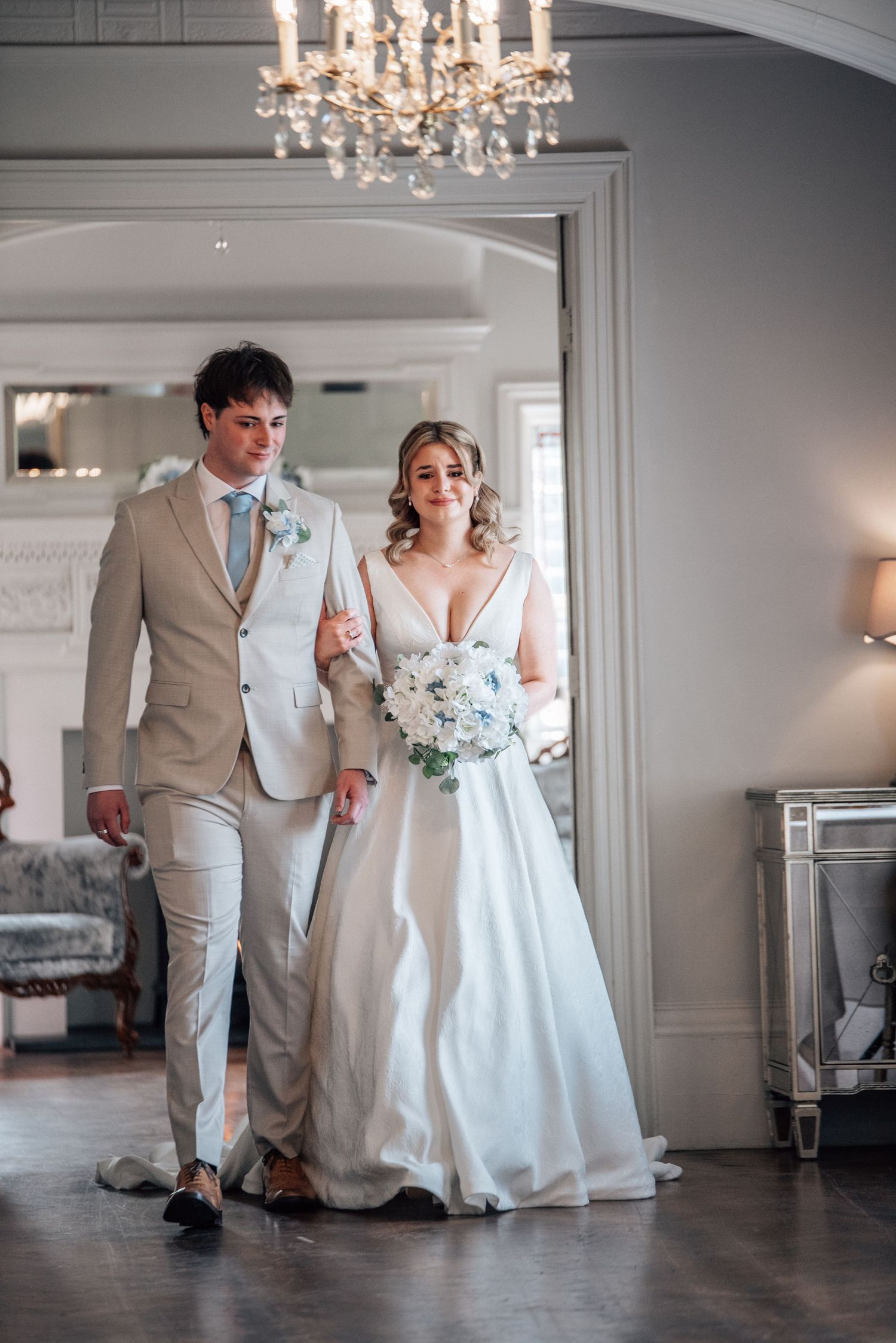 Bride and groom walking arm in arm through elegant venue doorway, bride holding blue and white bouquet, groom in beige suit.