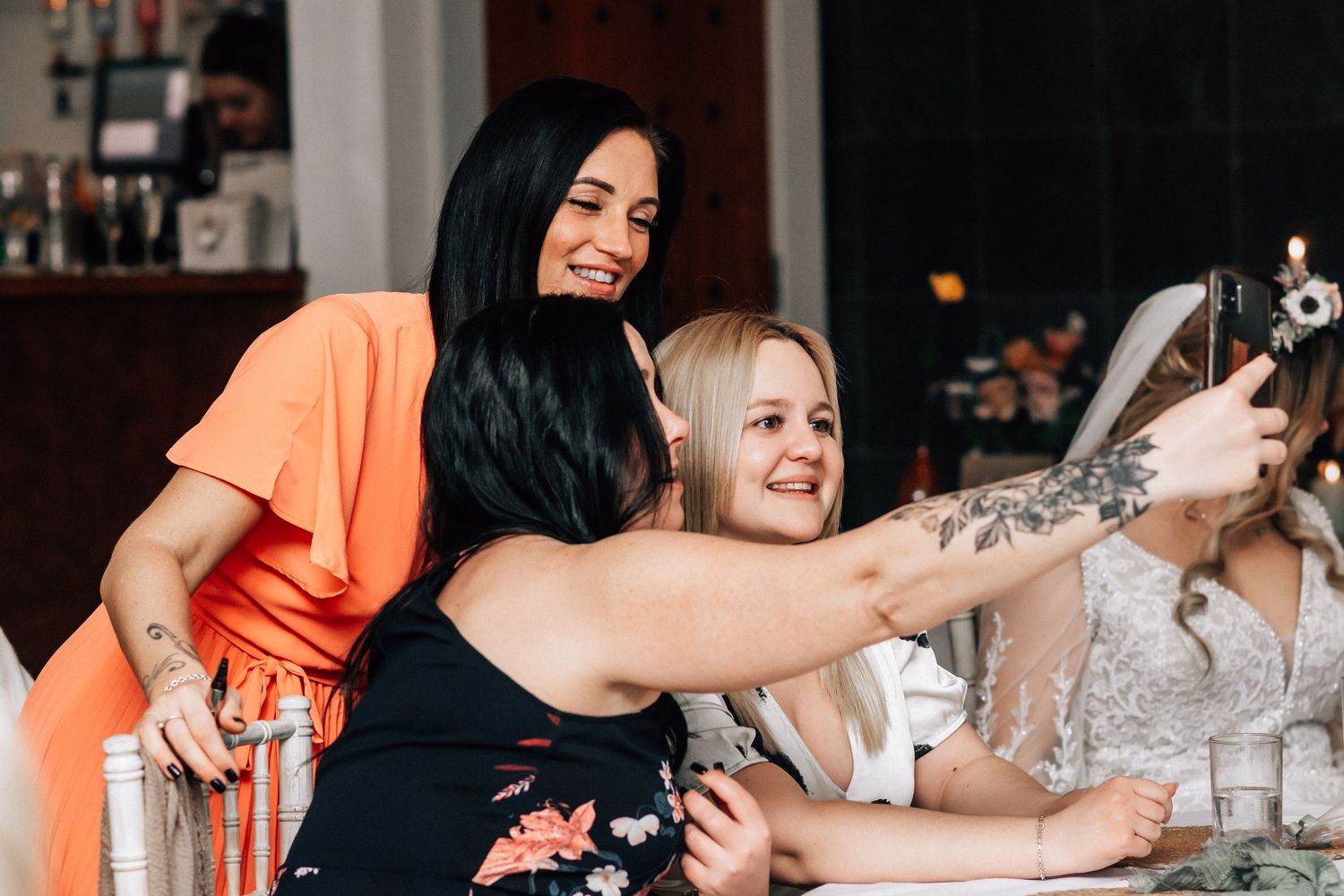 Three women taking a selfie together at a wedding reception, one wearing an orange top and another in a floral dress.