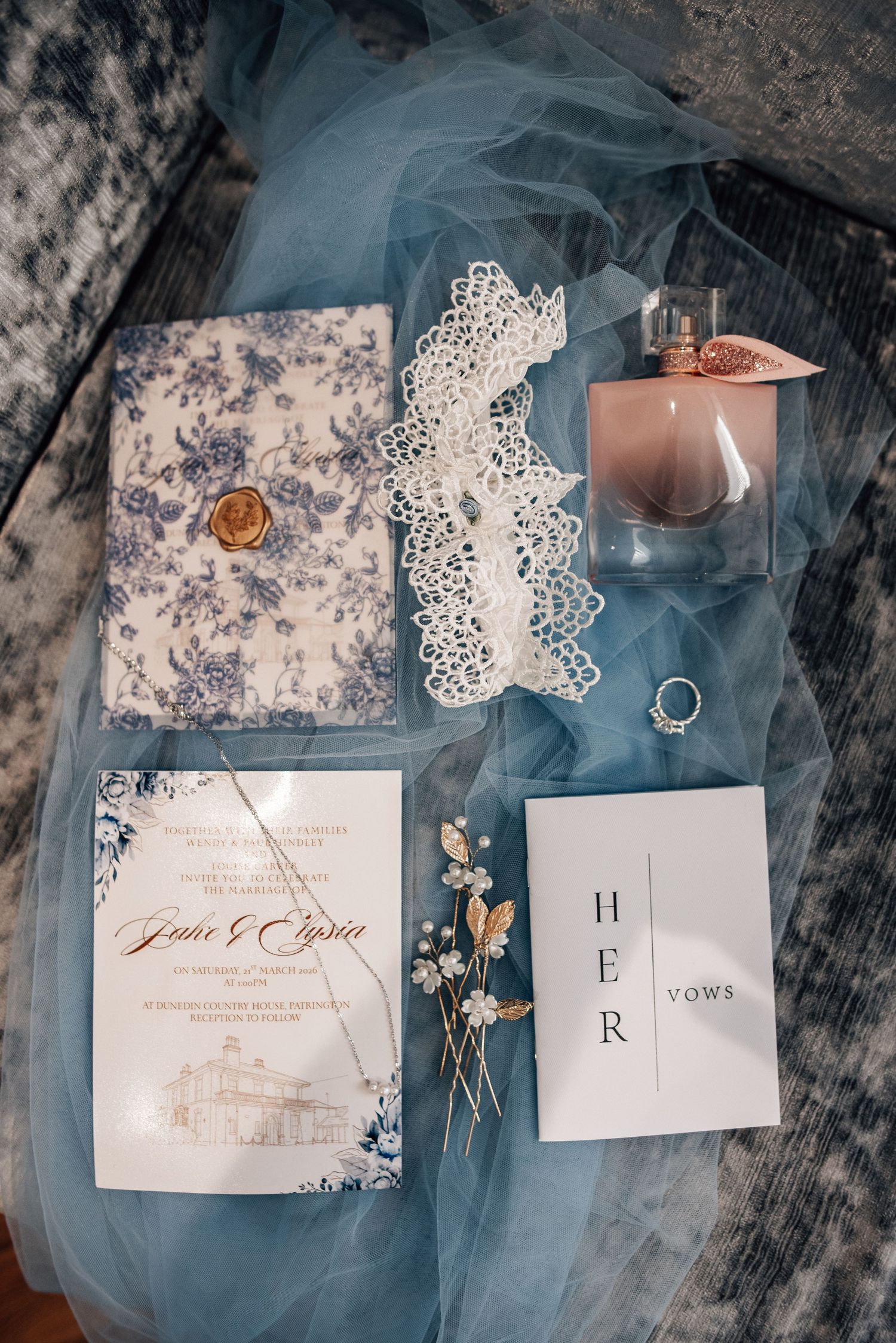 Elegant wedding flat lay with blue floral invitation suite, vow booklet, perfume, lace garter, and dried florals on dusty blue tulle.