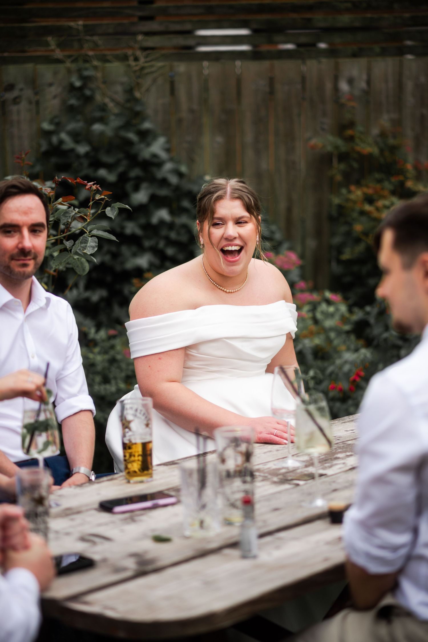 Joyful bride in off-shoulder white gown laughing with guests at outdoor wedding reception table.