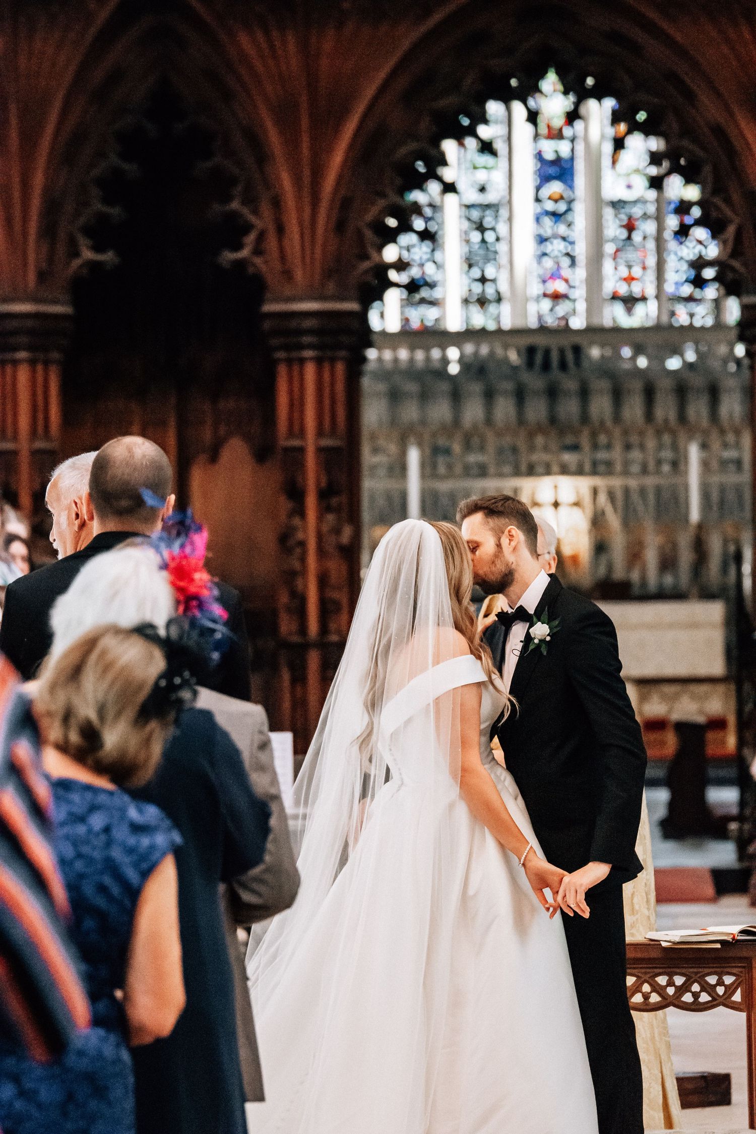 Bride and groom share first kiss at church altar with stained glass windows during wedding ceremony.