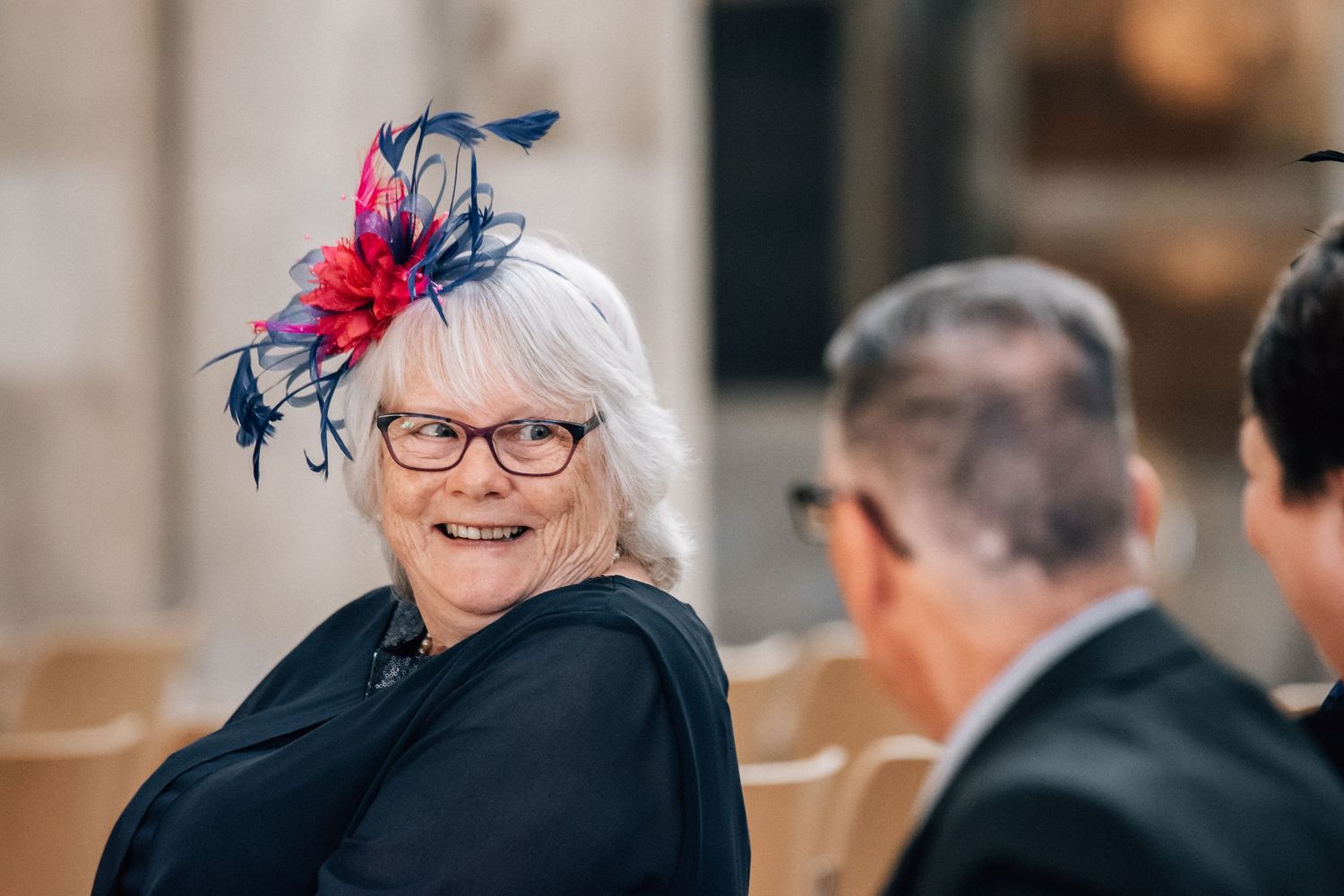 Smiling elderly woman with white hair, red glasses, and colorful fascinator hat at a formal event.