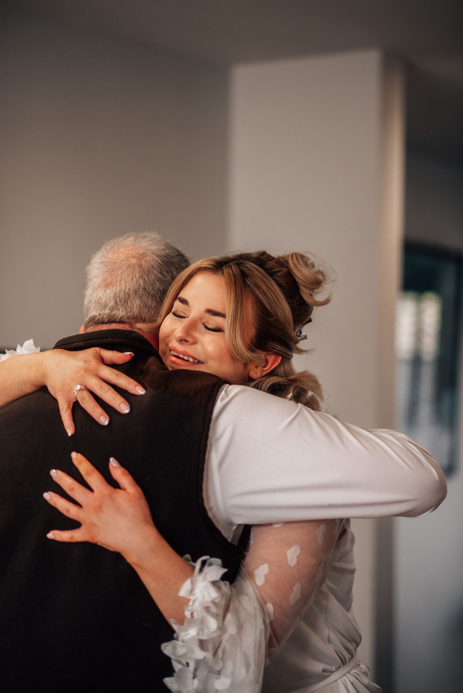 A smiling bride in white hugs an older man in a dark suit during an emotional wedding day moment.
