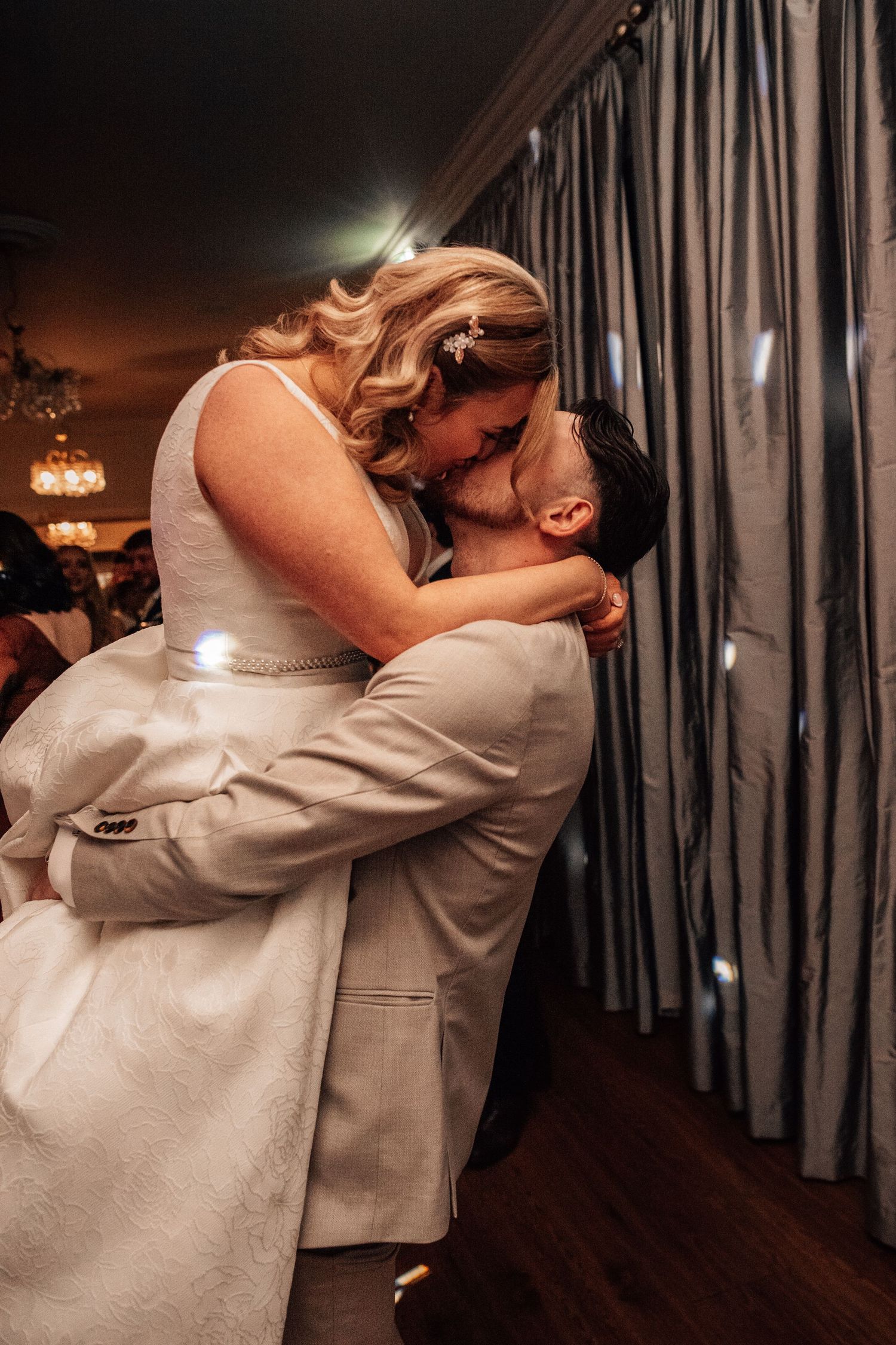 Groom lifting bride for a romantic kiss at their wedding reception near gray curtains.