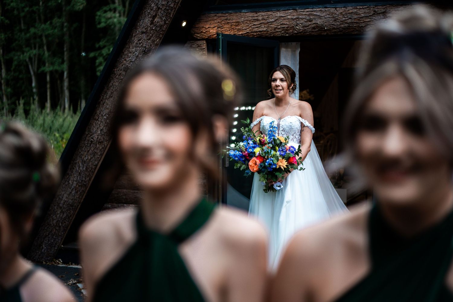 Bride in white gown holding colorful bouquet reflected in mirror, with blurred bridesmaids in green dresses in foreground.