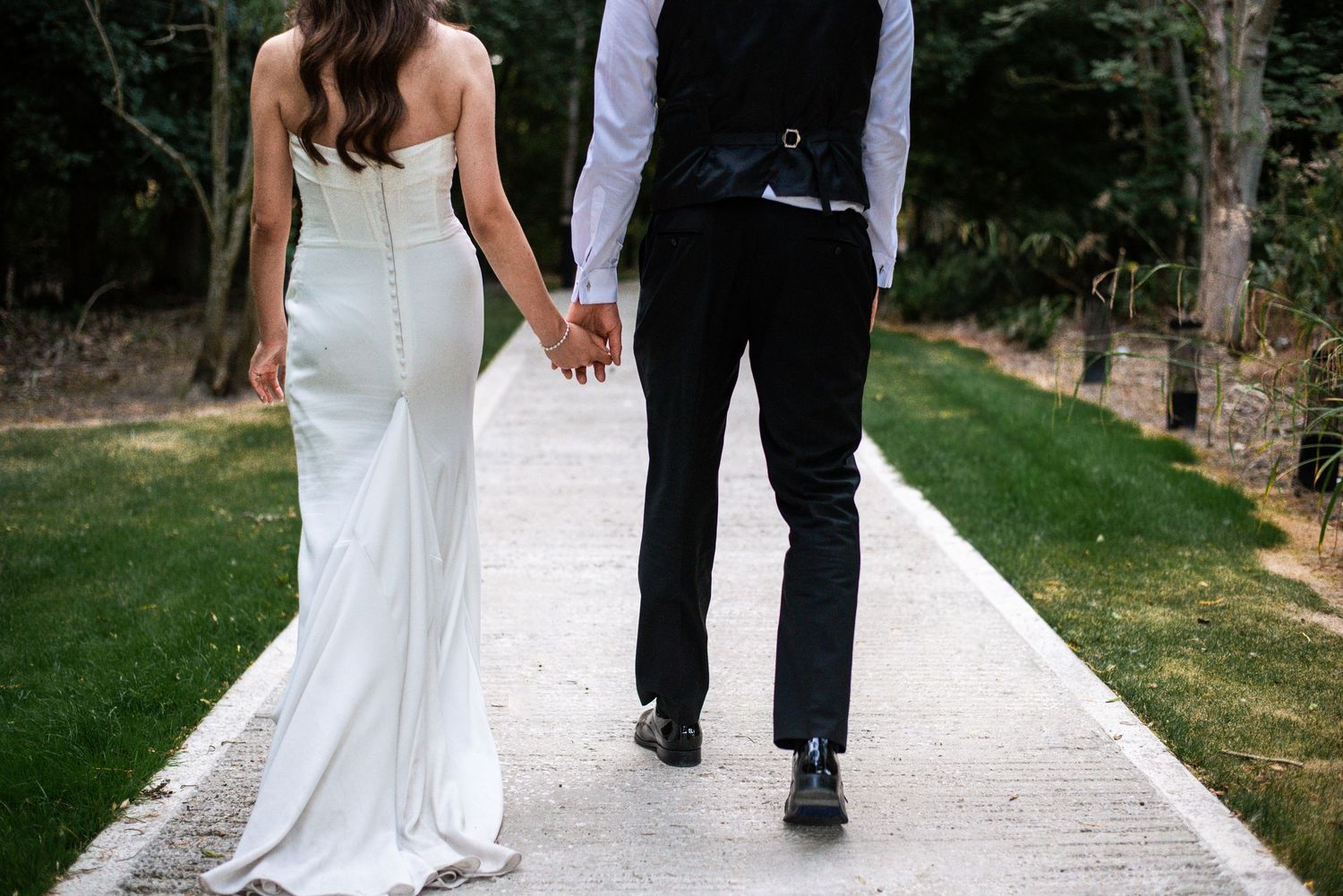 Bride and groom walking hand in hand down a garden path, viewed from behind on their wedding day.
