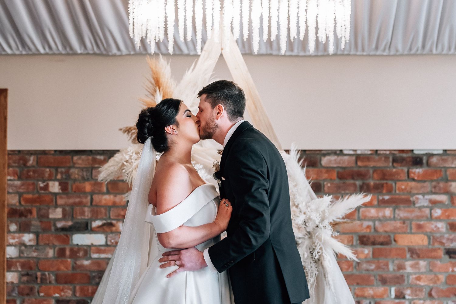 Bride and groom sharing a first kiss at boho wedding ceremony with teepee backdrop and exposed brick wall.