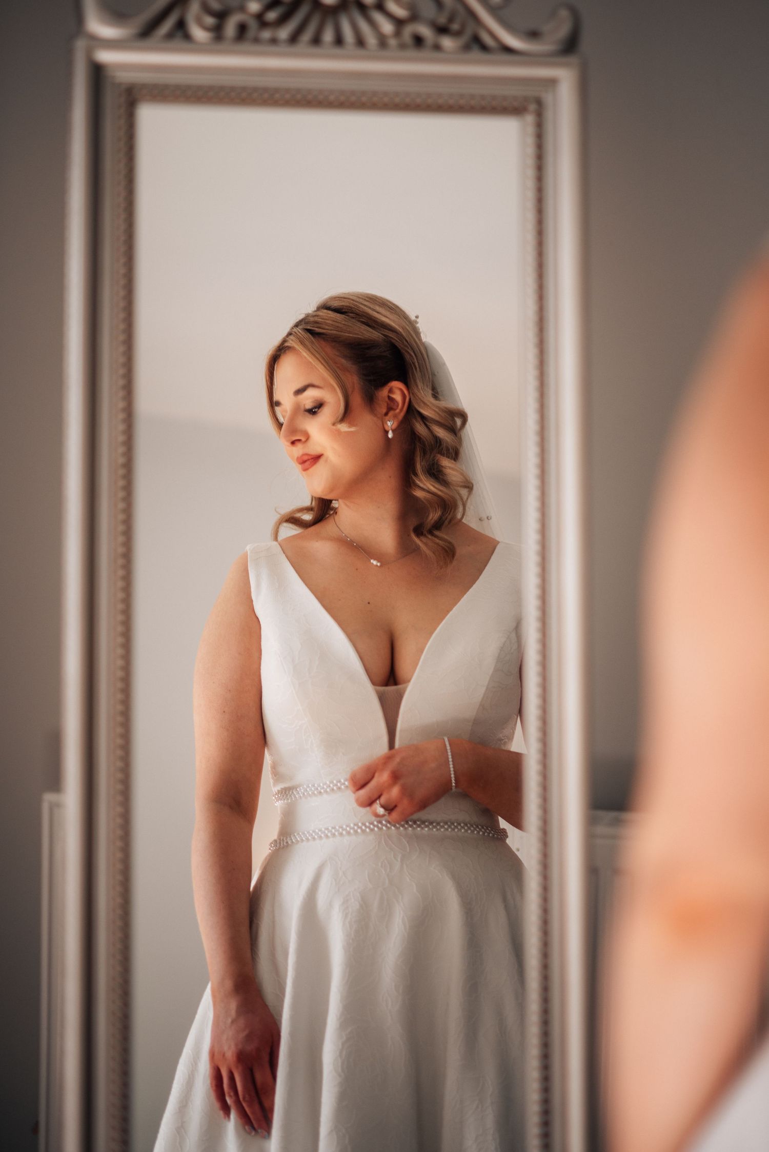 Elegant bride in a deep V-neck white wedding dress admiring herself in an ornate mirror.