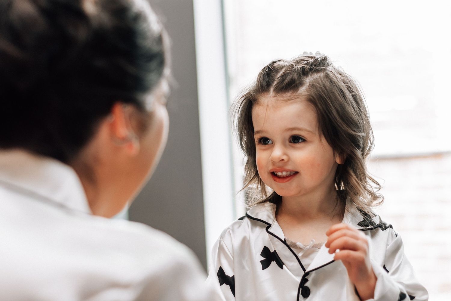 A smiling little girl in a white pajama set with bow print looks up at a woman in a bright indoor setting.