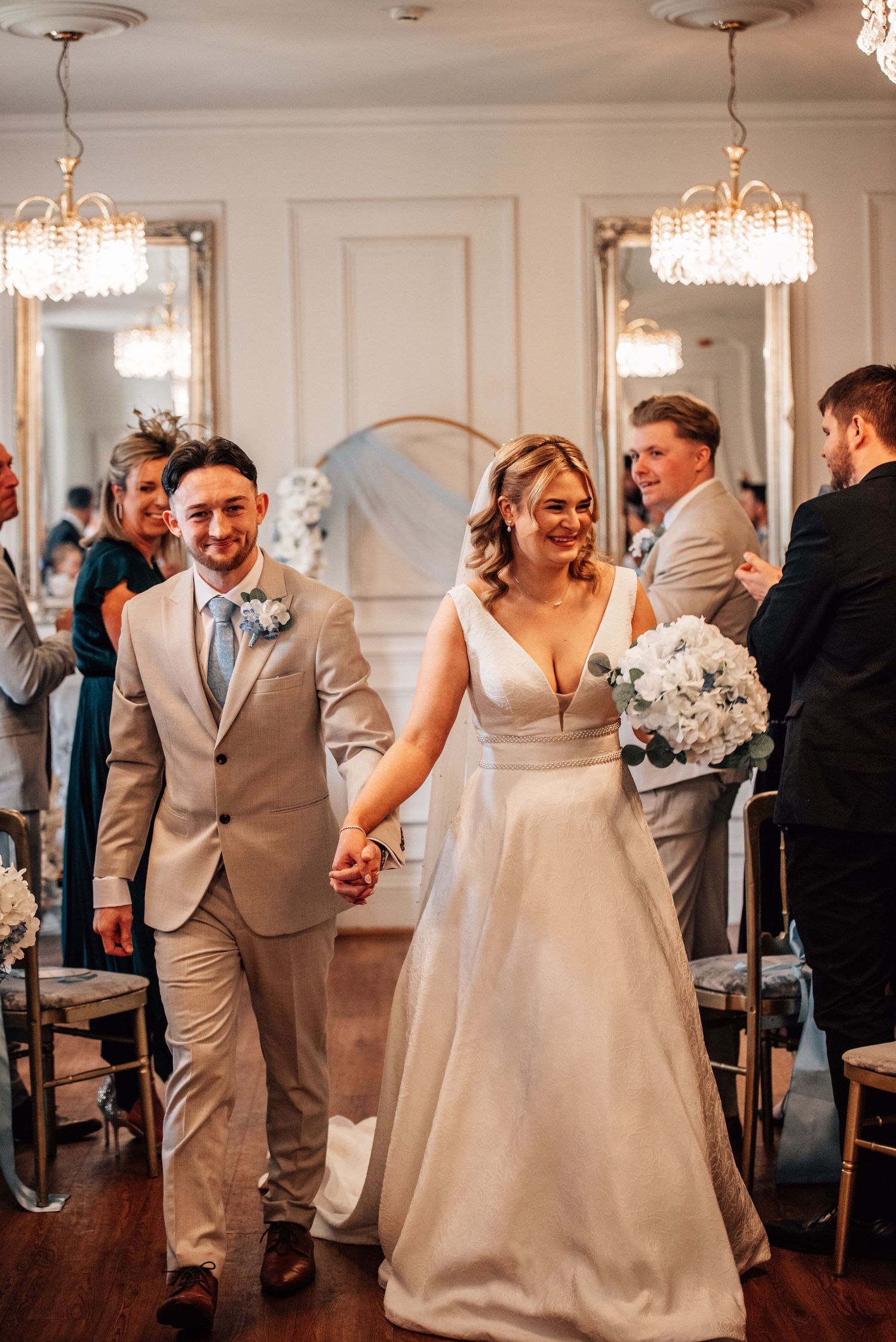 Smiling newlyweds walk hand-in-hand down the aisle after their elegant indoor wedding ceremony with crystal chandeliers.