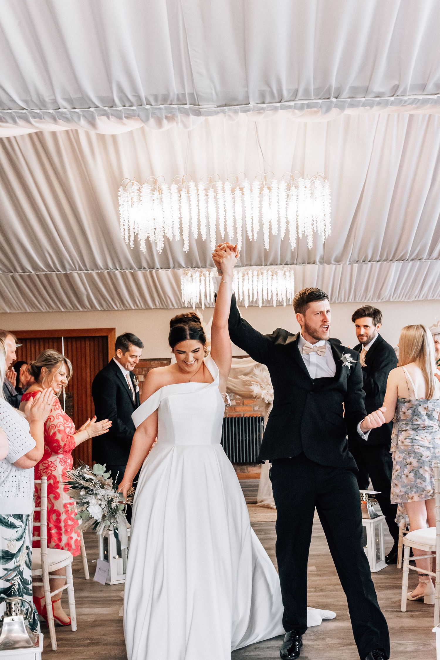 Bride and groom celebrate their first dance, raising hands joyfully in an elegant white draped wedding venue.