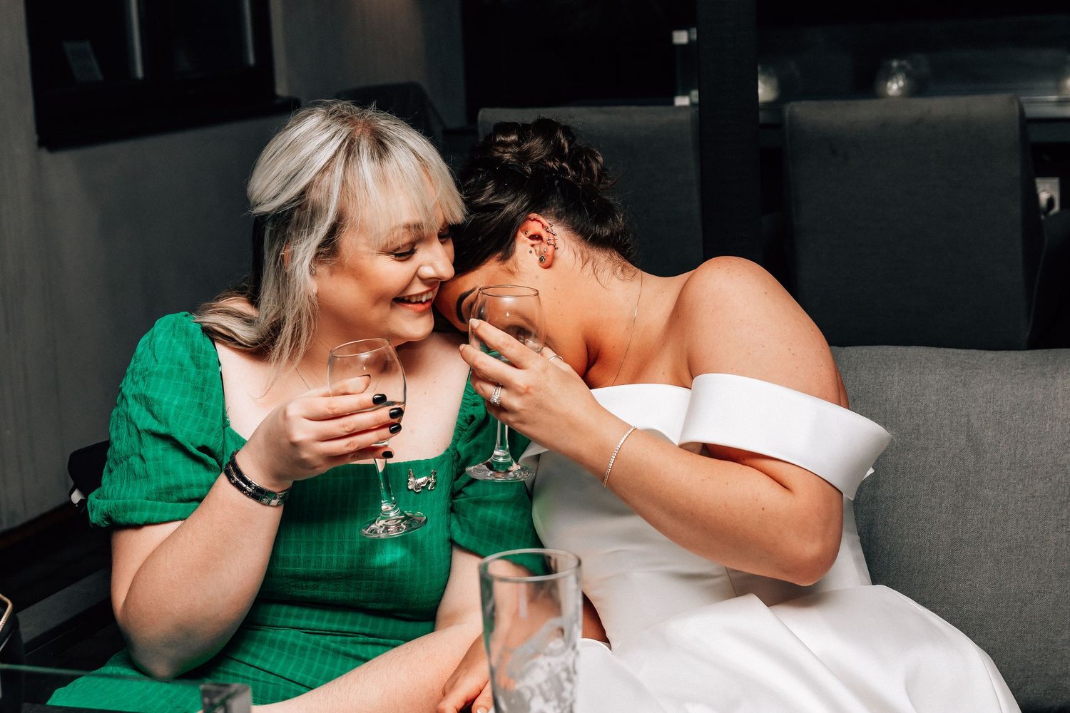 Bride in white off-shoulder gown shares an intimate moment with woman in green dress at wedding reception.