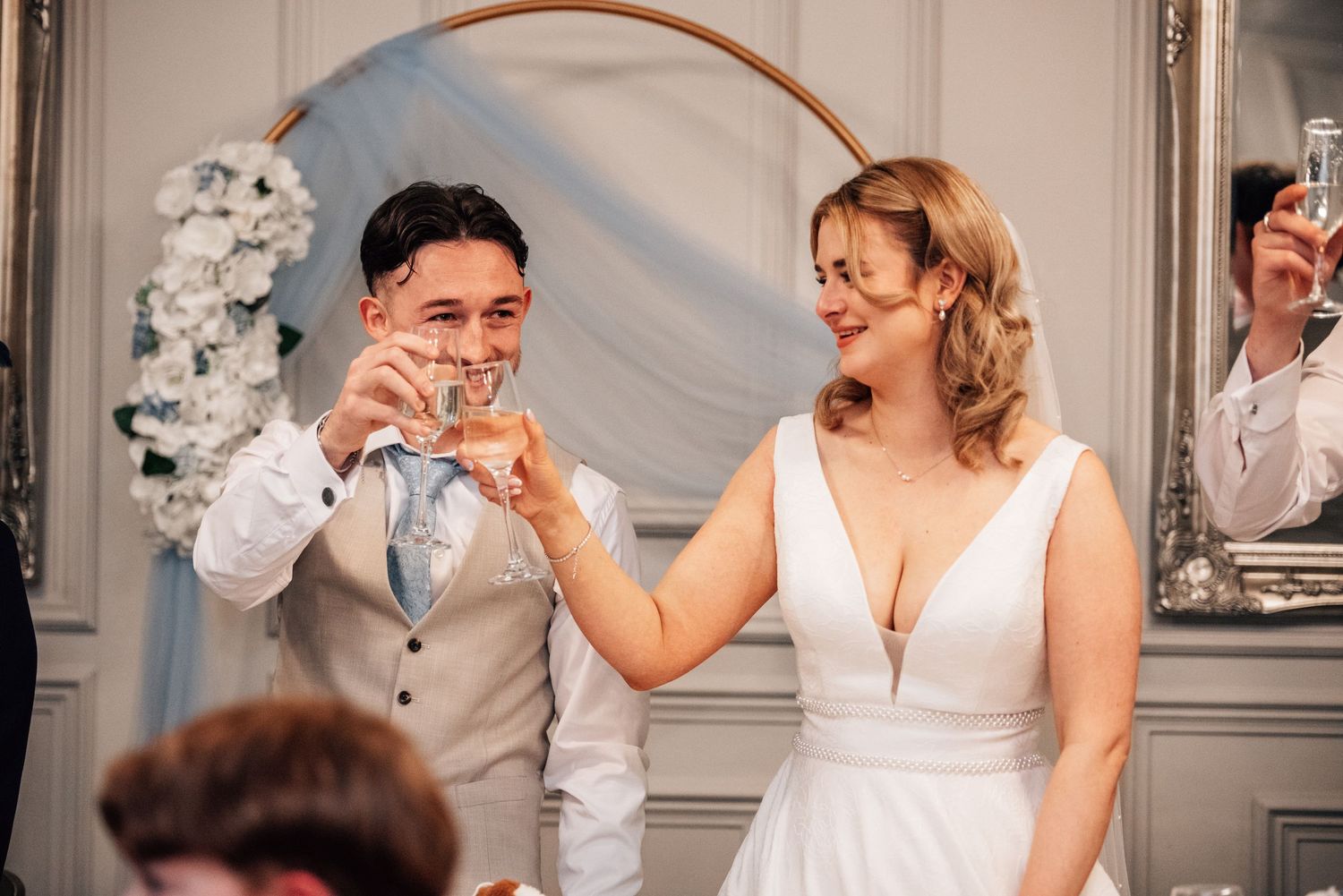 Bride and groom toasting with champagne glasses at their wedding reception near a floral arch backdrop.