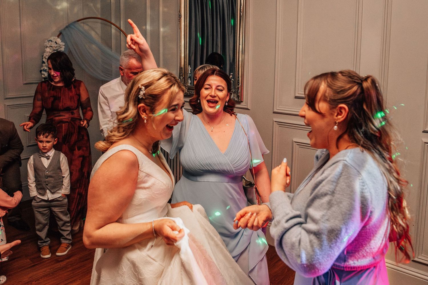 Bride laughing and dancing with guests at wedding reception party under colorful lights in elegant venue.