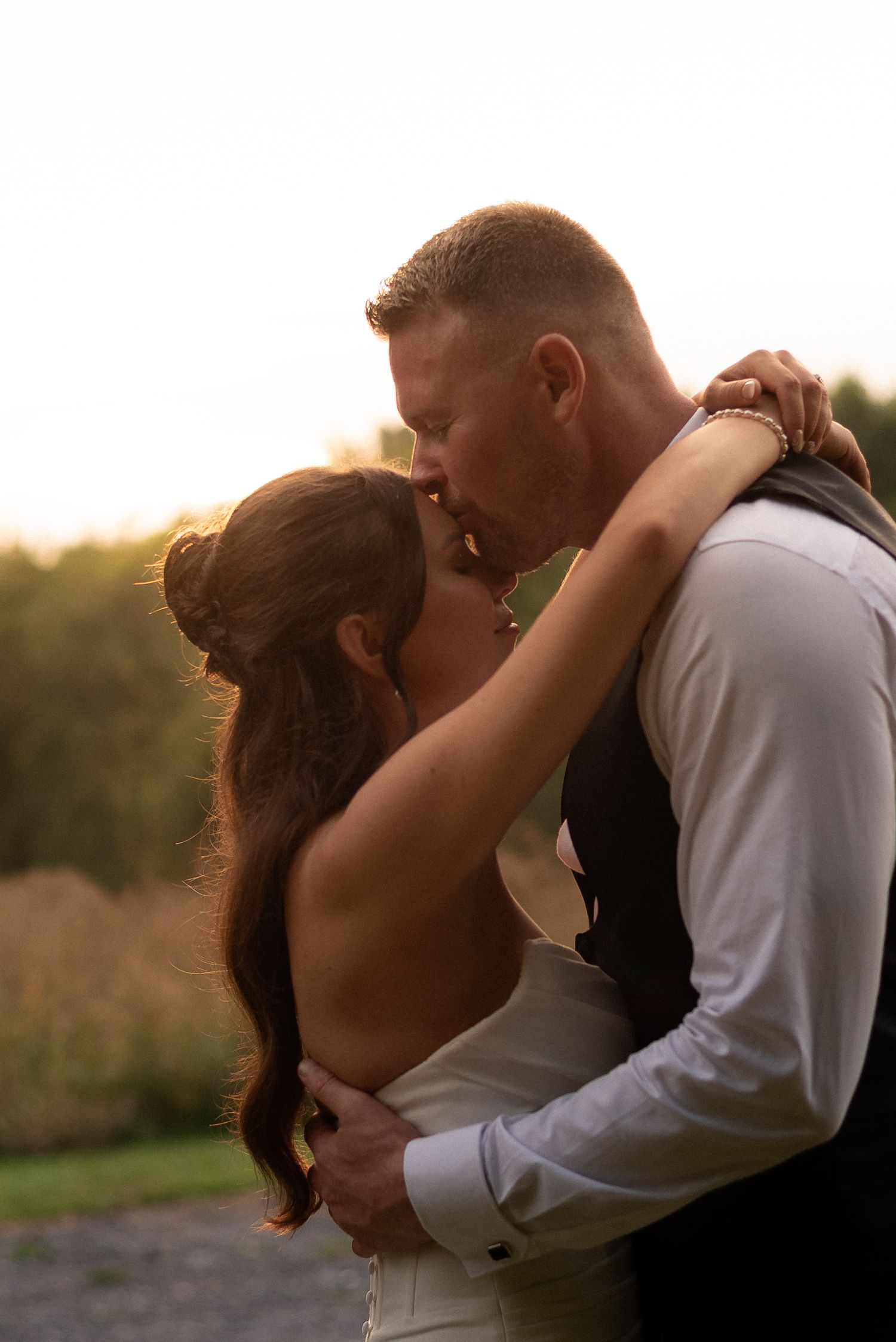A bride and groom share a romantic kiss at golden hour, embraced outdoors with warm sunset light glowing behind them.