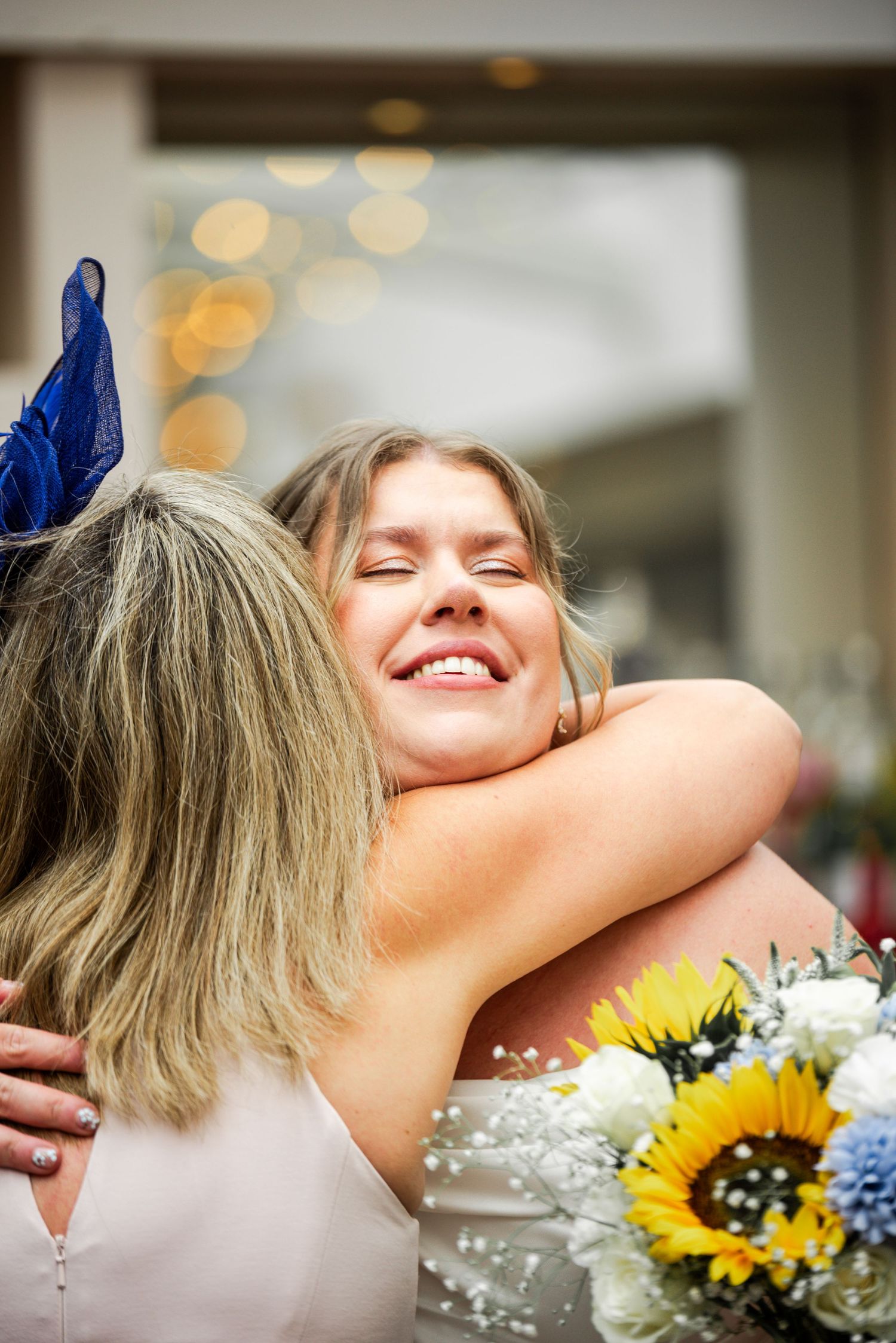 Smiling bride hugging a guest wearing a blue fascinator while holding a sunflower bouquet.
