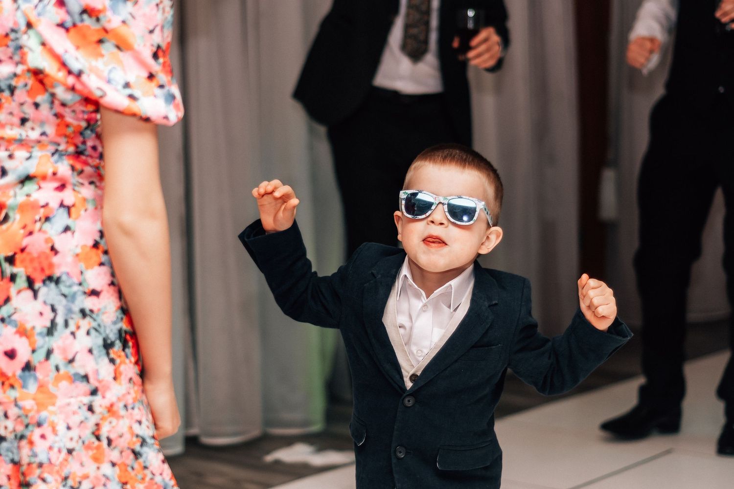 A young boy in a navy suit and white sunglasses dances joyfully at a wedding reception.