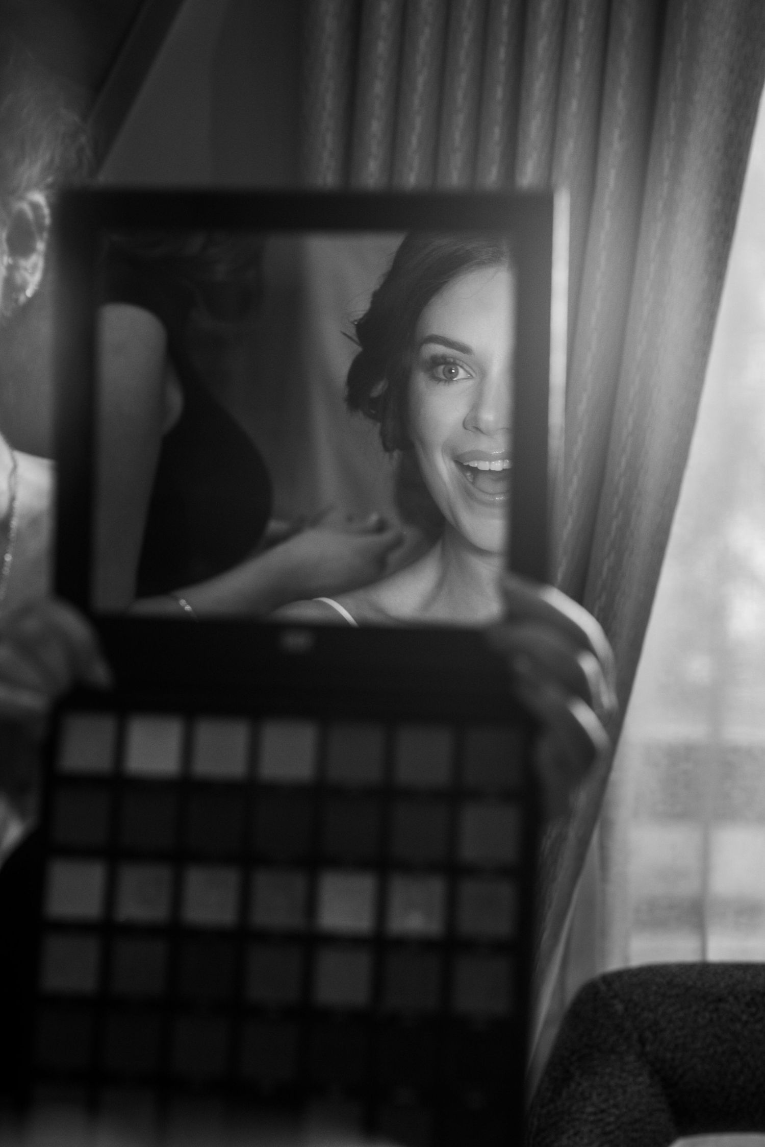 Black and white photo of a smiling bride seeing her makeup look in a mirror held by her makeup artist.