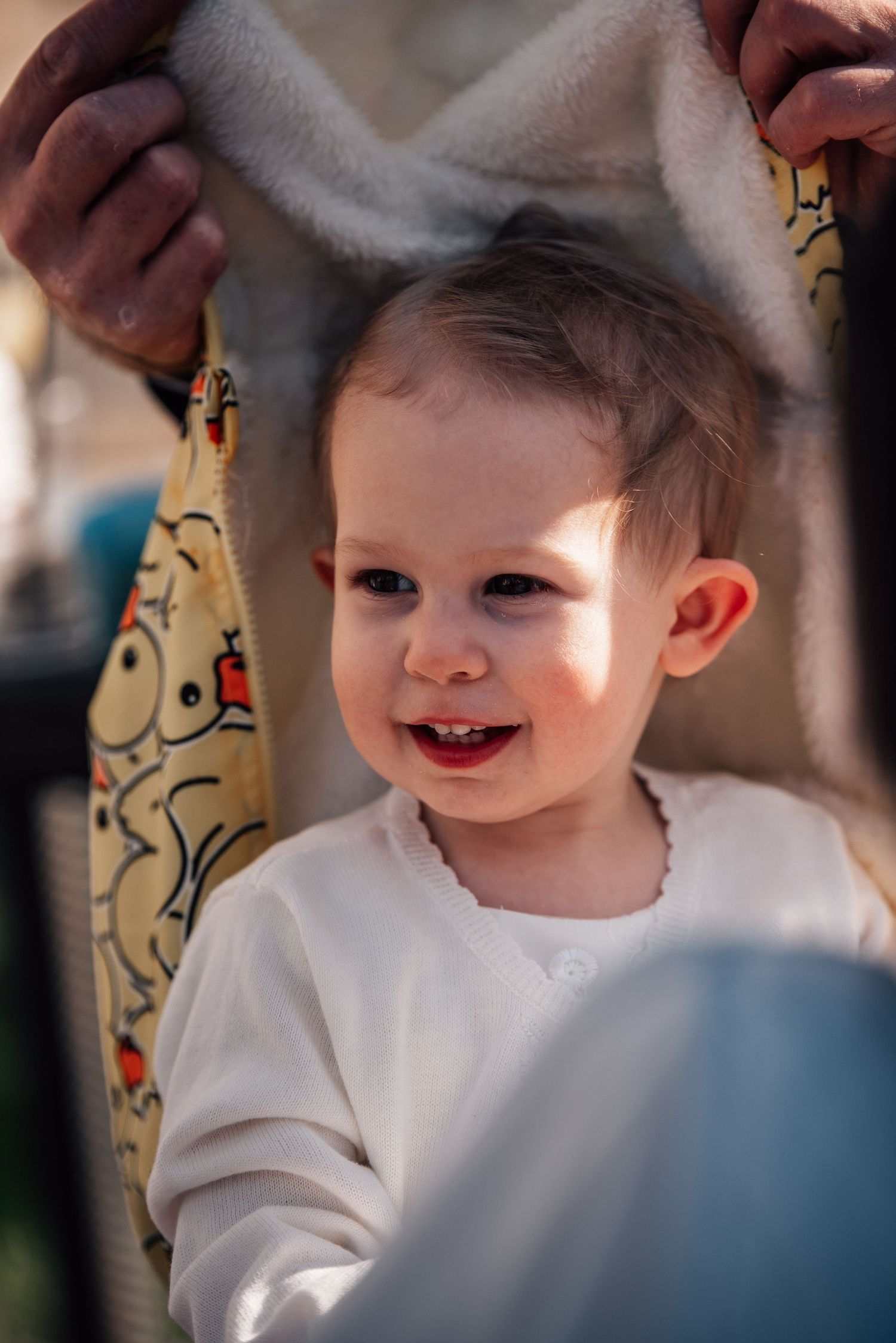 Happy smiling toddler in white outfit being playfully covered with a colorful blanket outdoors.