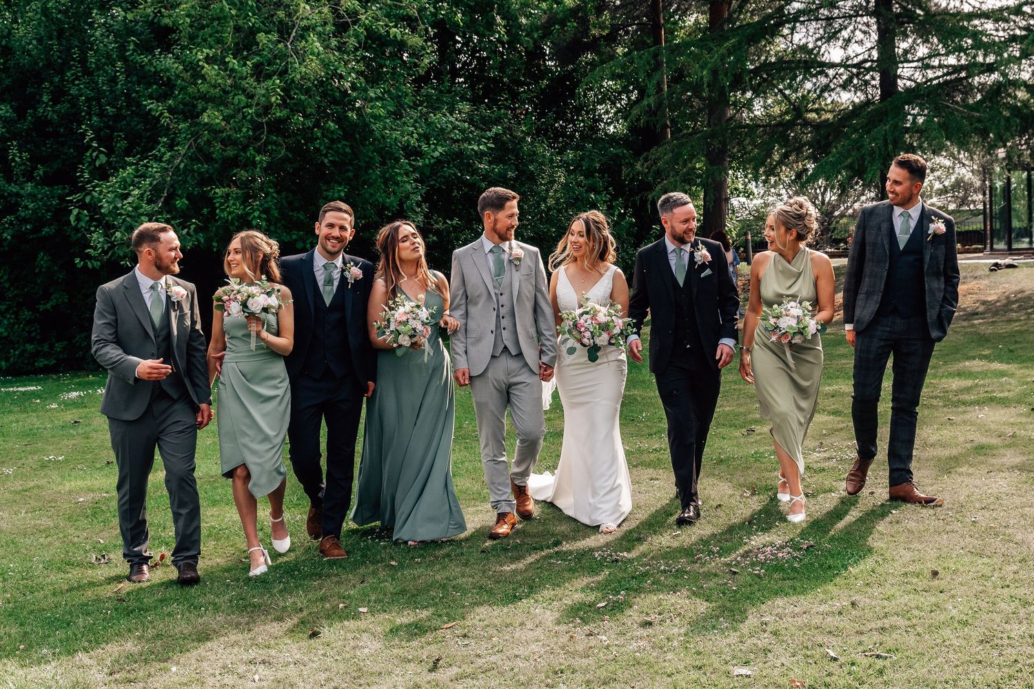 Wedding party walks outdoors on grass, bride in white gown with bridesmaids in sage green and groomsmen in suits.