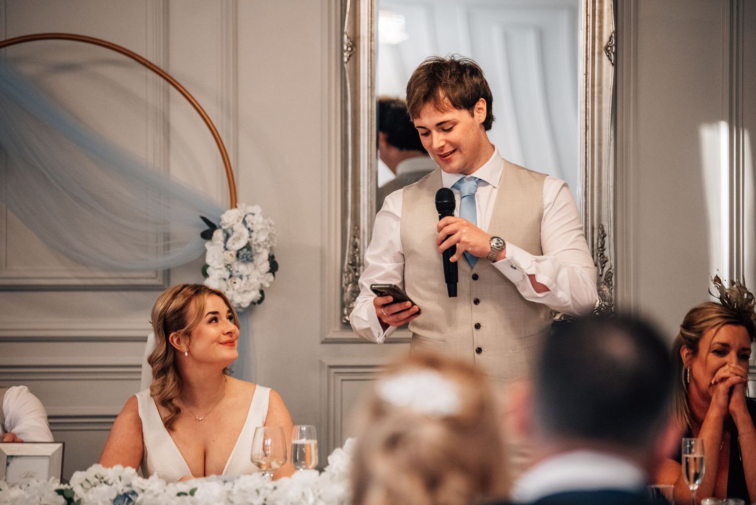 Groom in a beige suit delivering a wedding speech with microphone while bride smiles at the table.