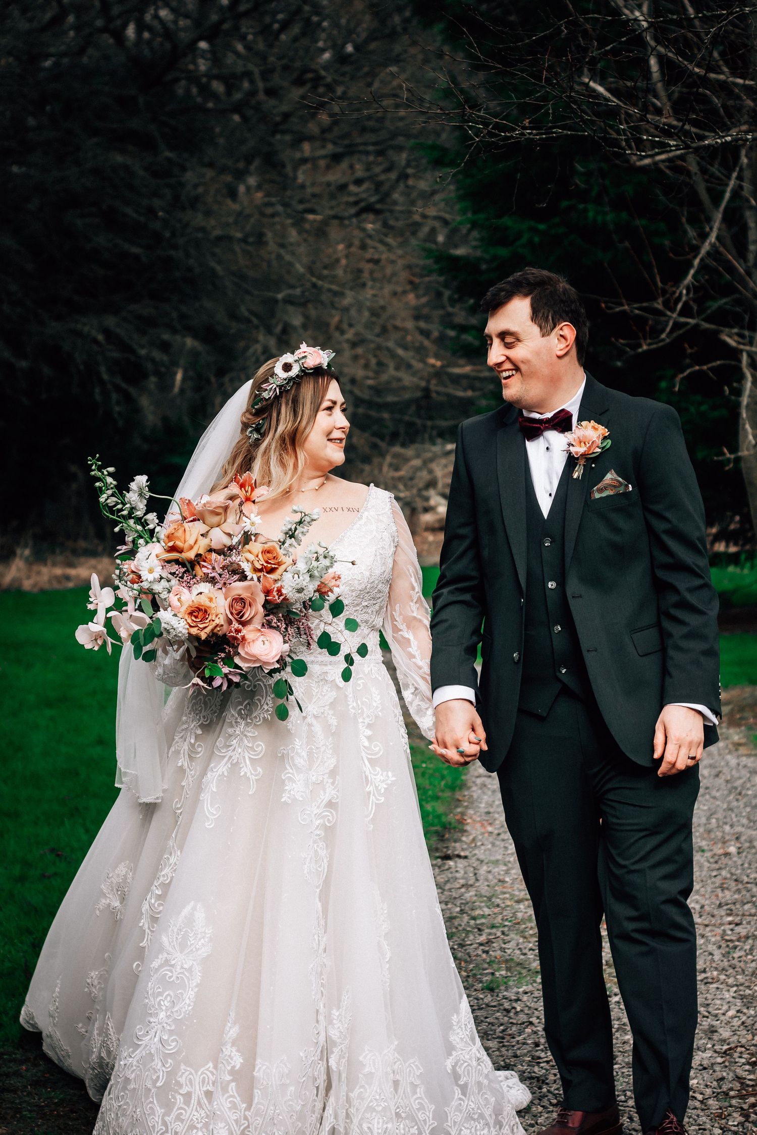 Bride and groom walk hand-in-hand, she holds a peach floral bouquet in a lace gown, he wears a dark green suit.