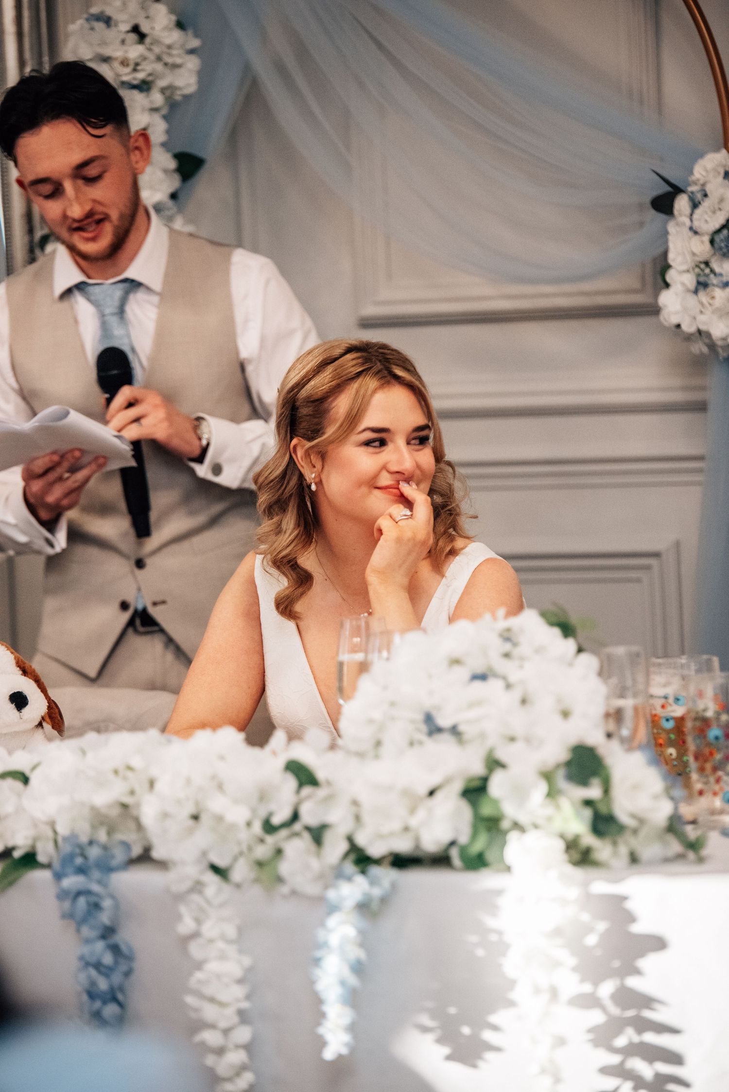 Bride smiling emotionally during wedding speech at elegant reception with white floral arrangements and blue draping.