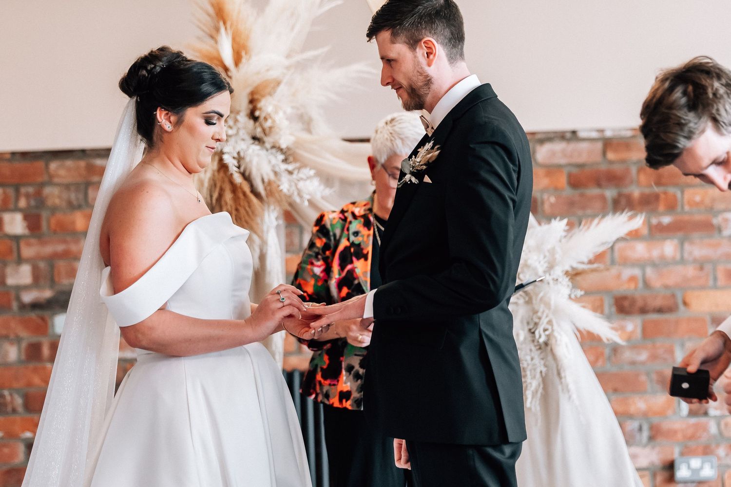 Bride and groom exchanging rings during wedding ceremony at brick wall venue.