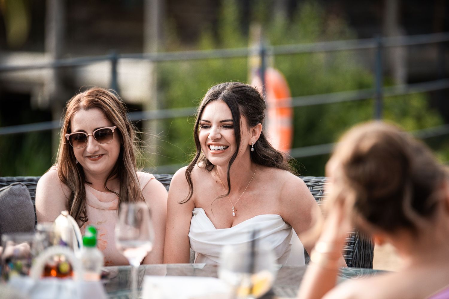 Two women smile and laugh at an outdoor wedding reception table, one wearing sunglasses, enjoying a sunny celebration.