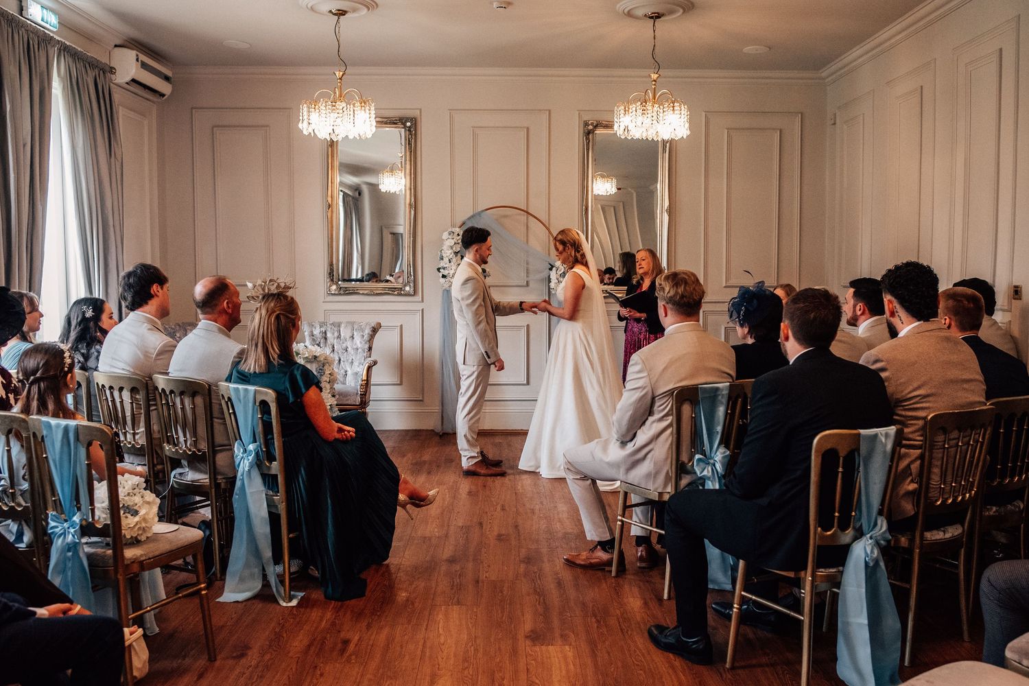 Couple exchanging vows at an elegant indoor wedding ceremony with guests seated on chairs adorned with teal ribbons.