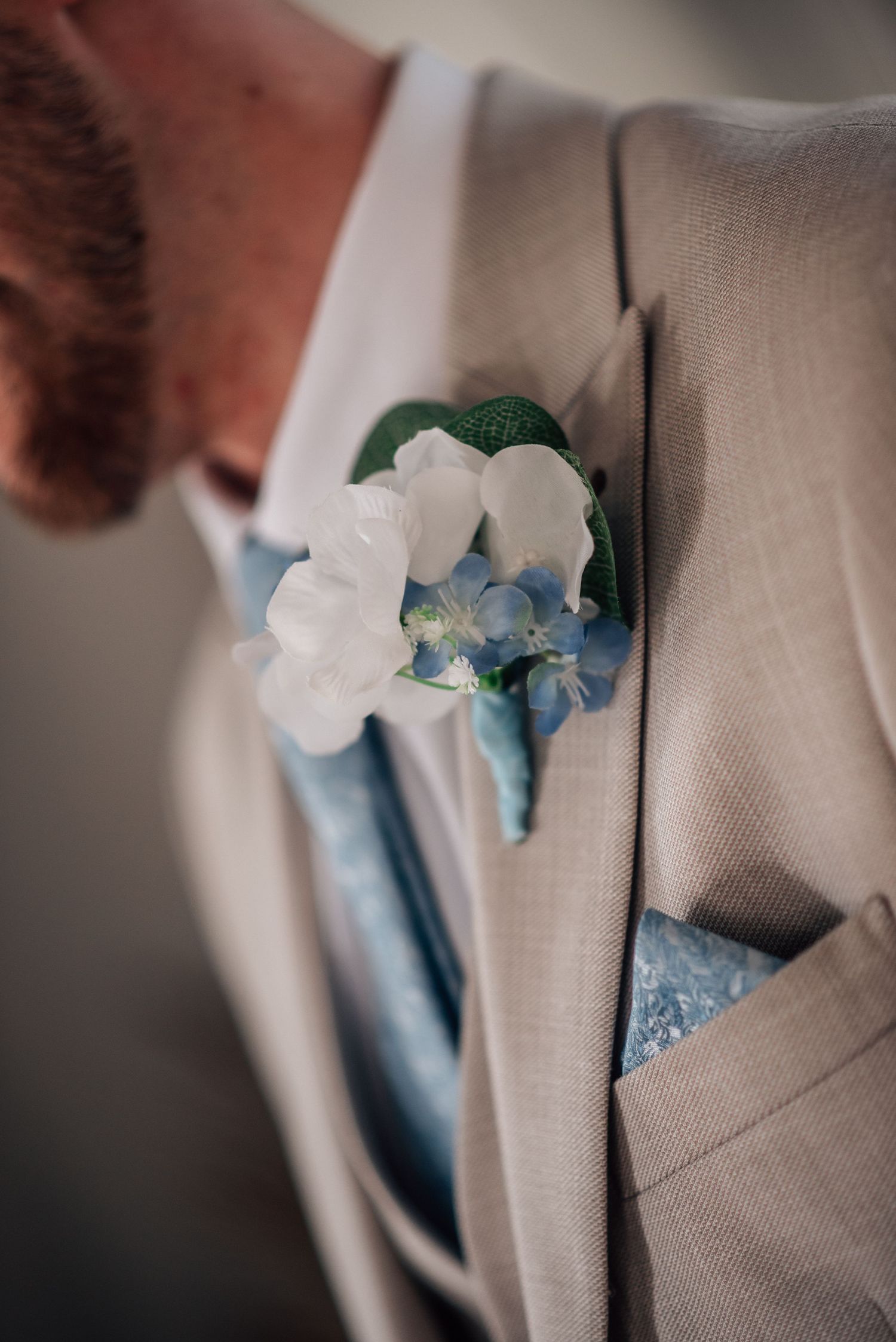 Close-up of a groom's beige suit lapel featuring a white and blue floral boutonniere with a matching pocket square.