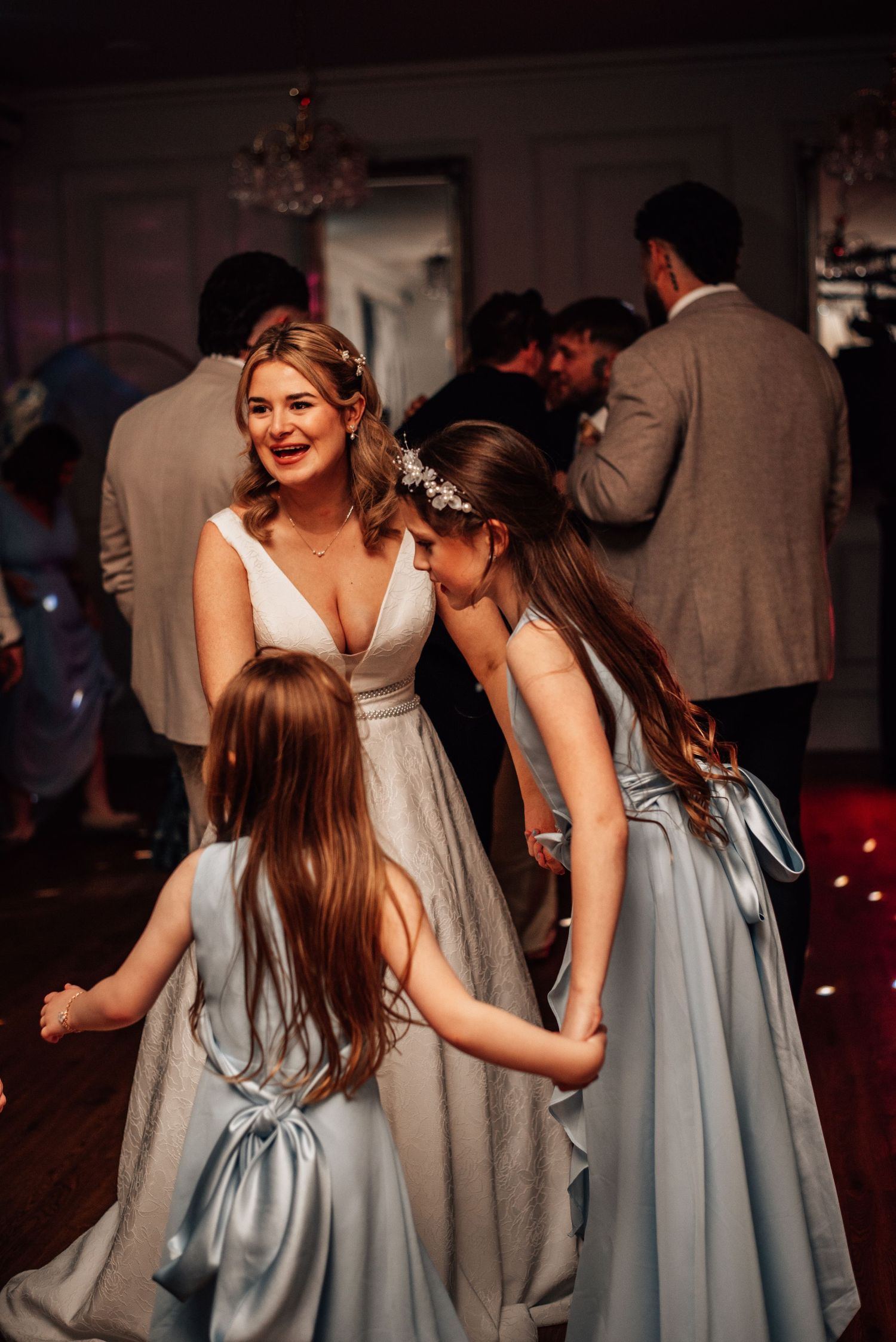 Smiling bride in white gown interacts warmly with two young flower girls in blue dresses on the dance floor.