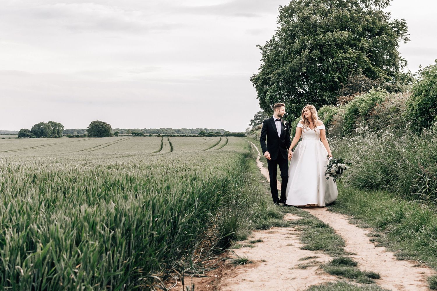 Bride and groom walk hand-in-hand along a countryside path beside green fields on their wedding day.