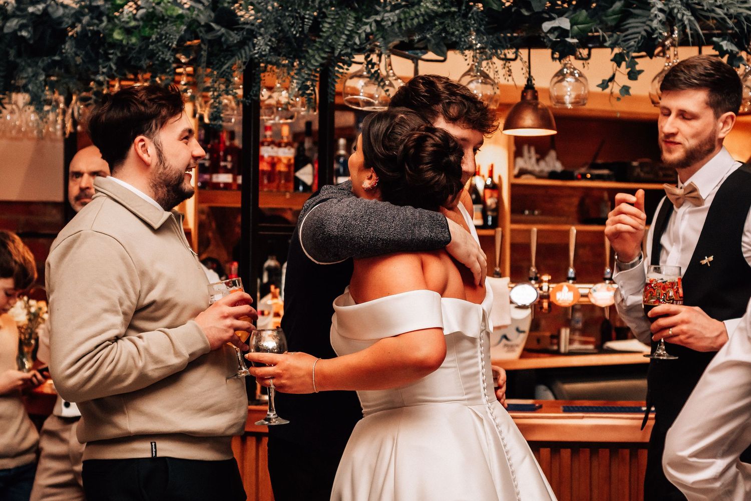 Bride in off-shoulder white gown shares a warm hug with a guest near a decorated bar.