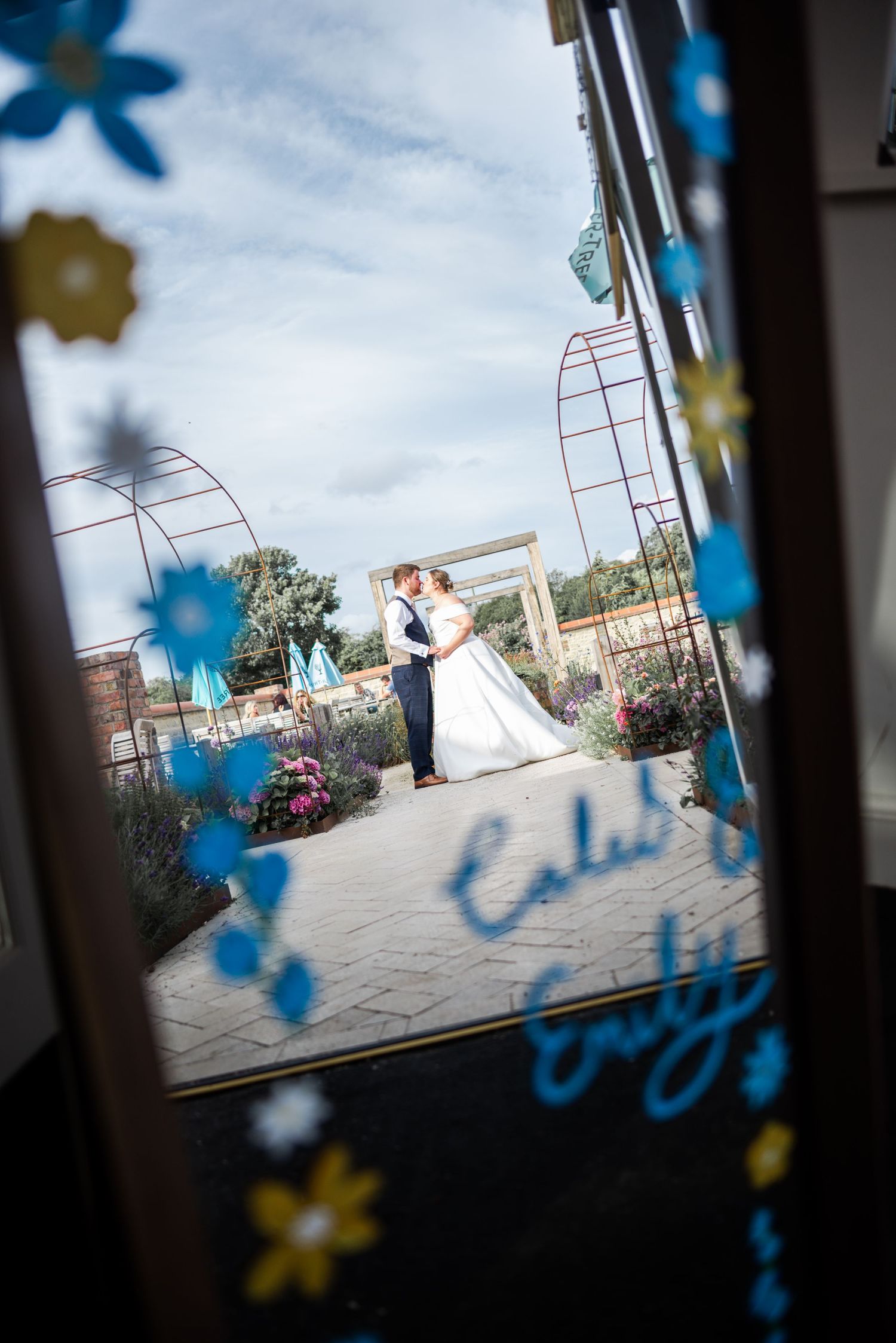 Bride and groom share a romantic kiss in a garden, viewed through a decorated glass door with blue flowers.
