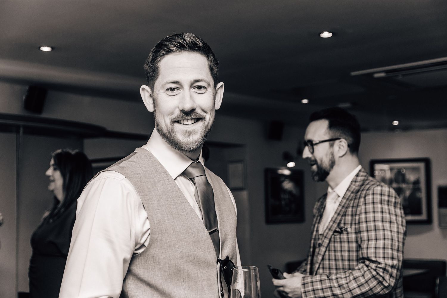 Smiling bearded man in waistcoat and tie at an indoor event in black and white photo.