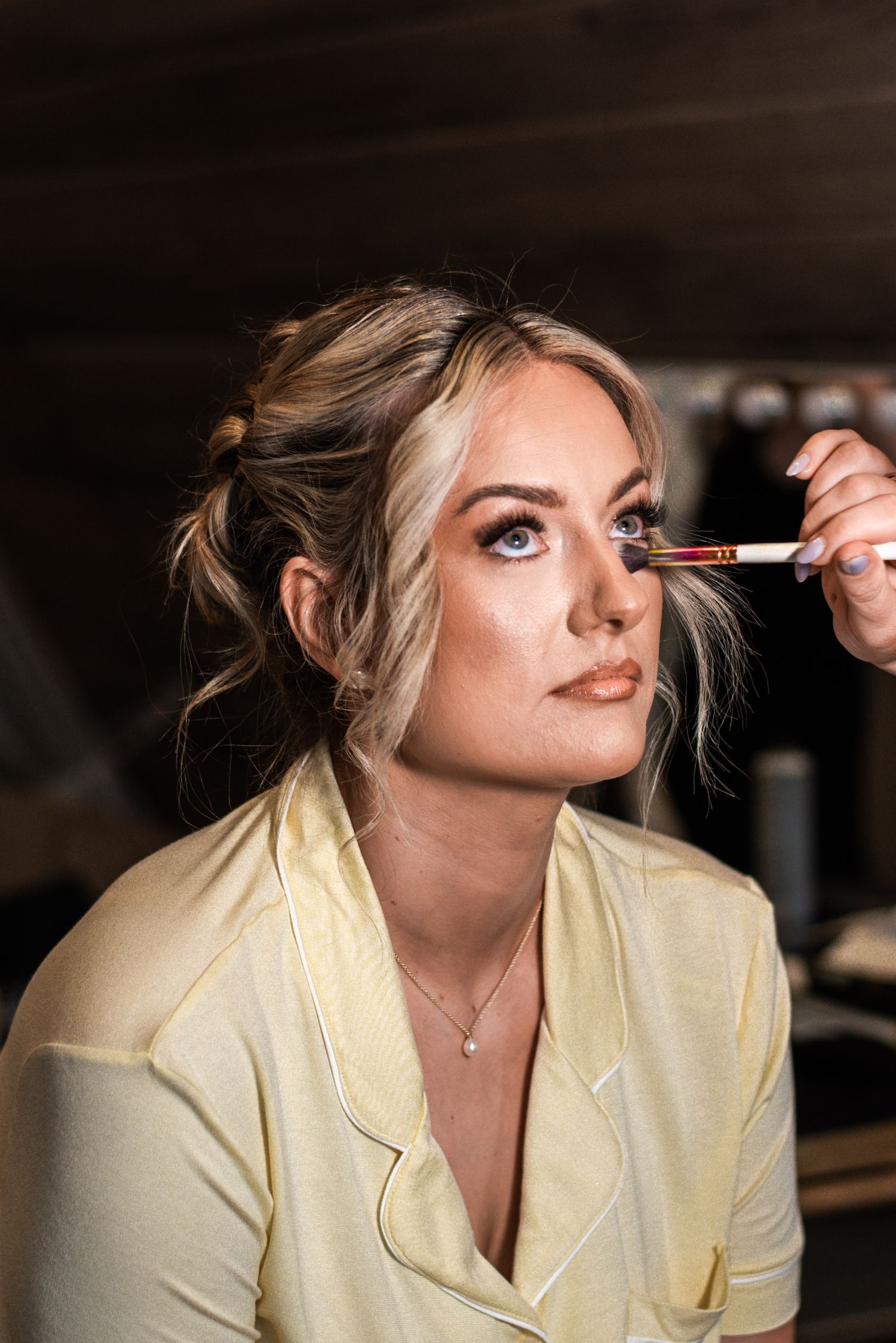 Blonde bride in yellow robe gets professional makeup applied with a brush before her wedding day.