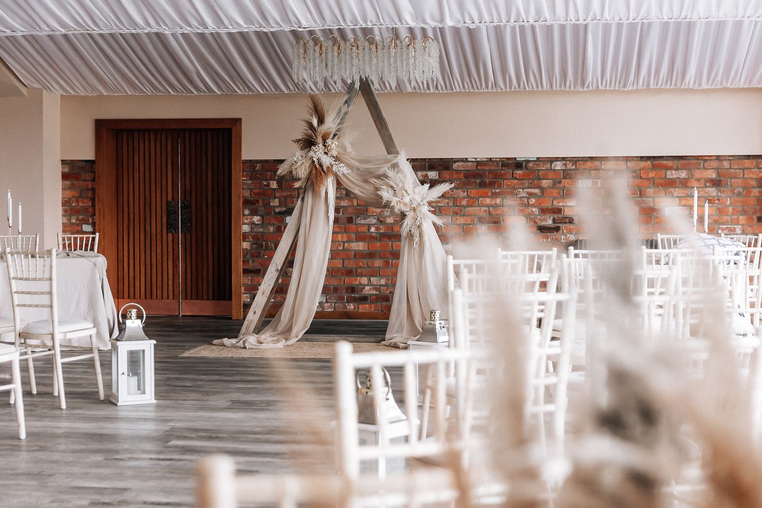 Elegant wedding ceremony venue with draped fabric arch, white Chiavari chairs, and exposed brick wall backdrop.
