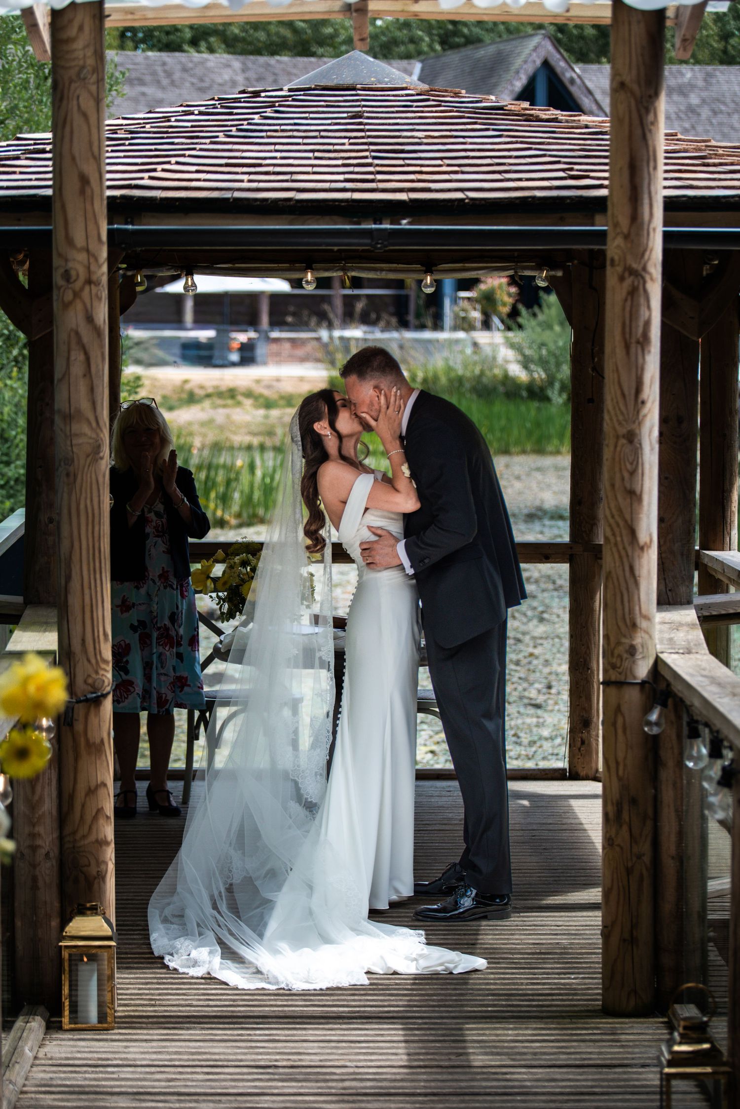 Bride and groom share first kiss under wooden gazebo during outdoor waterside wedding ceremony.