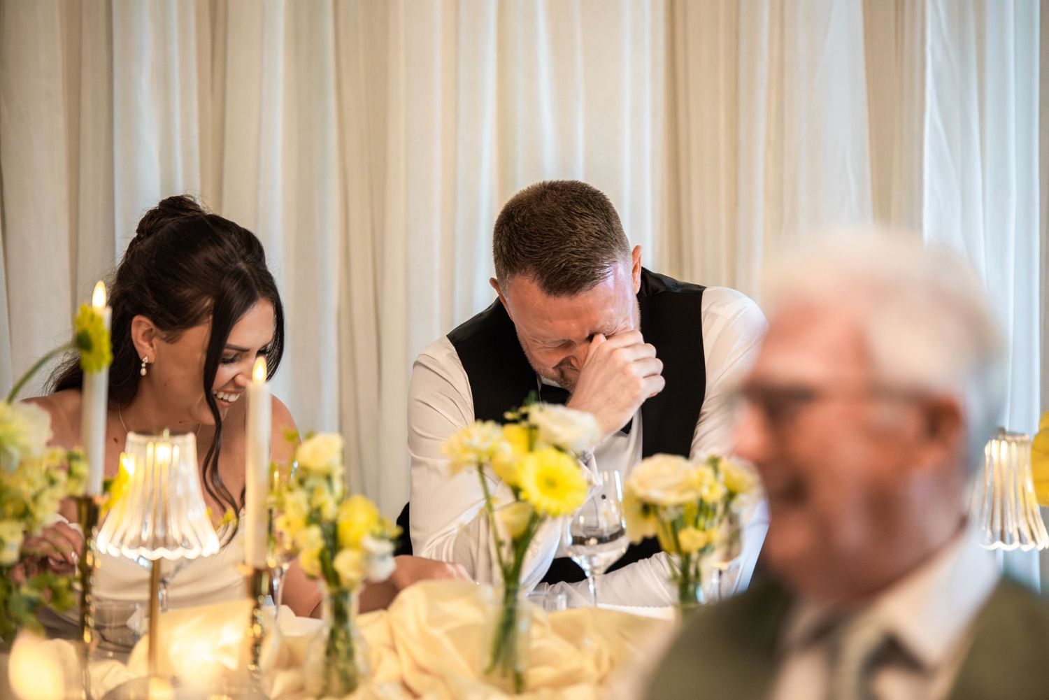 Bride and groom laughing together at wedding reception table adorned with yellow flowers and candles.