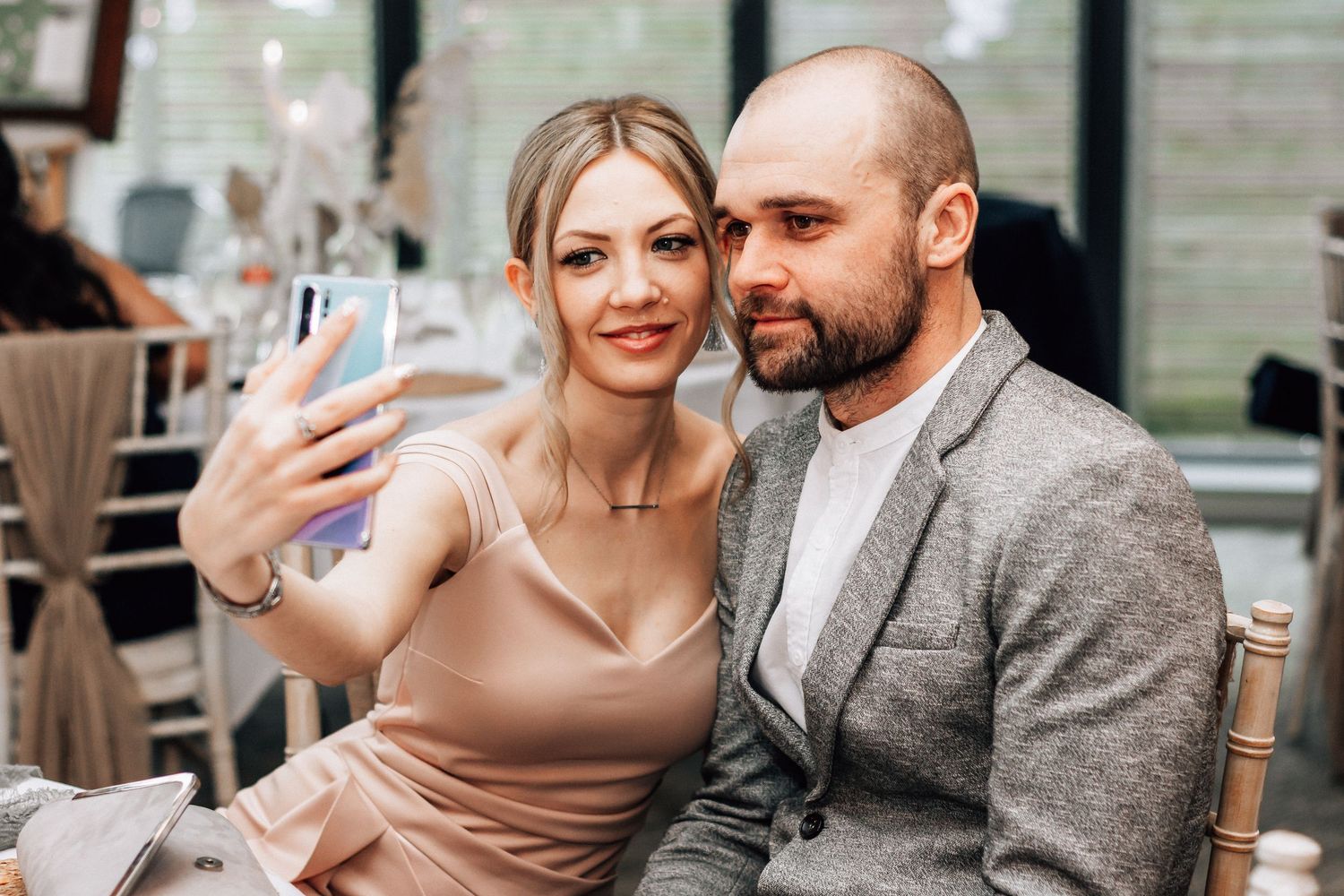 A smiling couple takes a selfie with a smartphone at an elegant event, woman in beige dress, man in gray blazer.