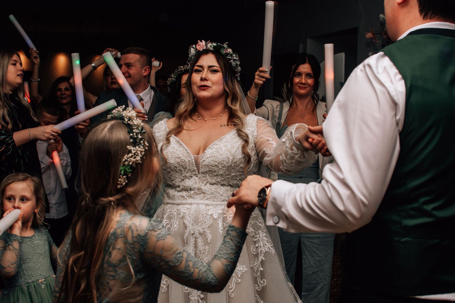Bride in lace gown with floral crown dances with guests holding glow sticks at a lively wedding reception.