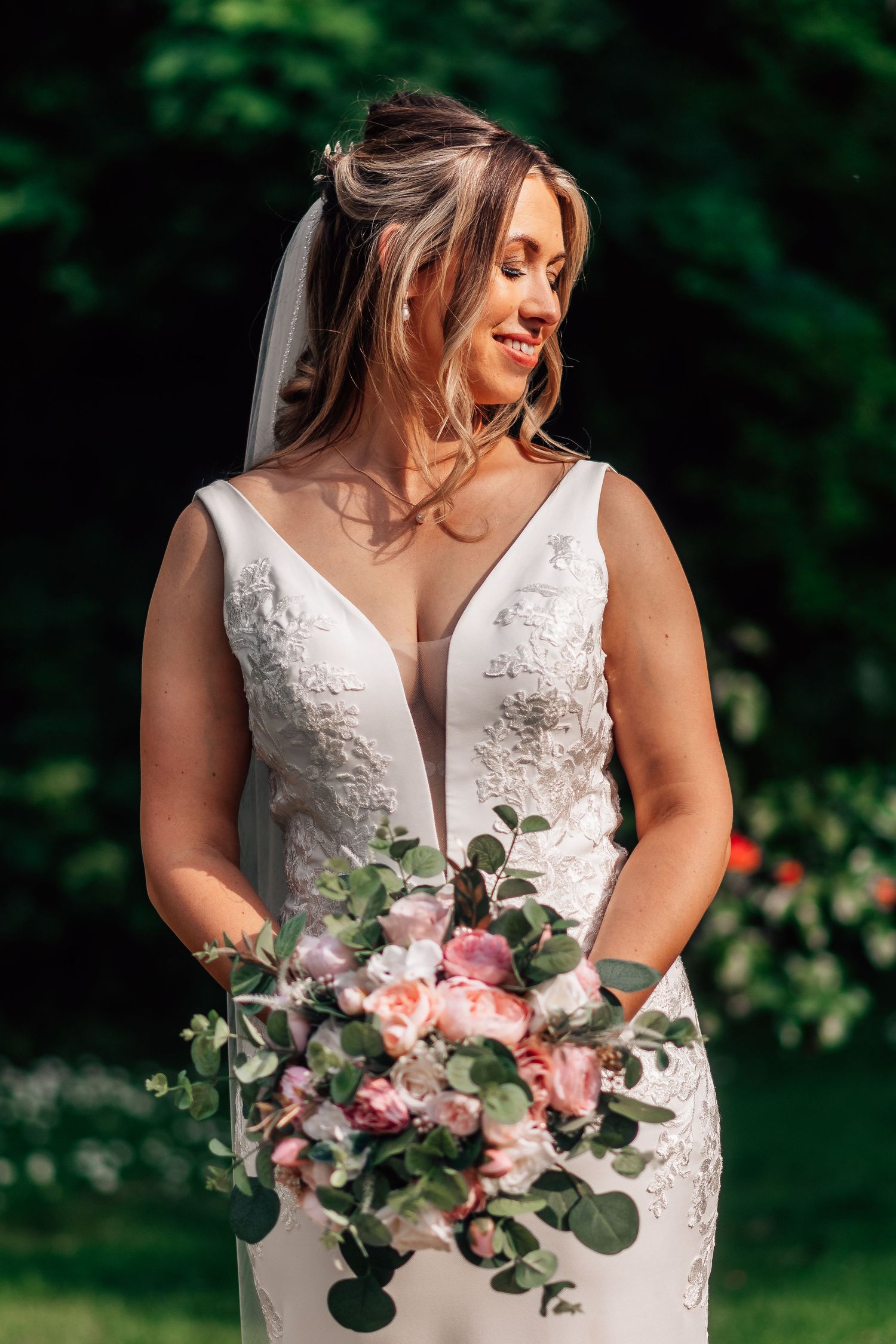 Beautiful bride in a deep V-neck lace wedding dress holding a cascading pink rose bouquet outdoors.
